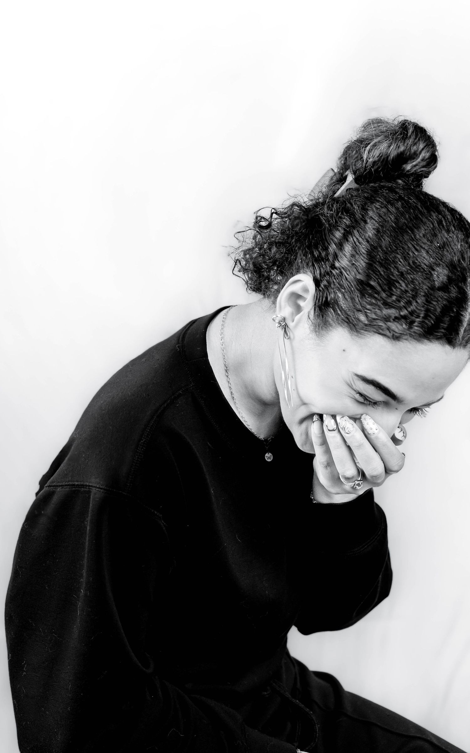 Black and white portrait of a young woman with curly hair in a bun, wearing a black sweatshirt, hands covering her mouth.