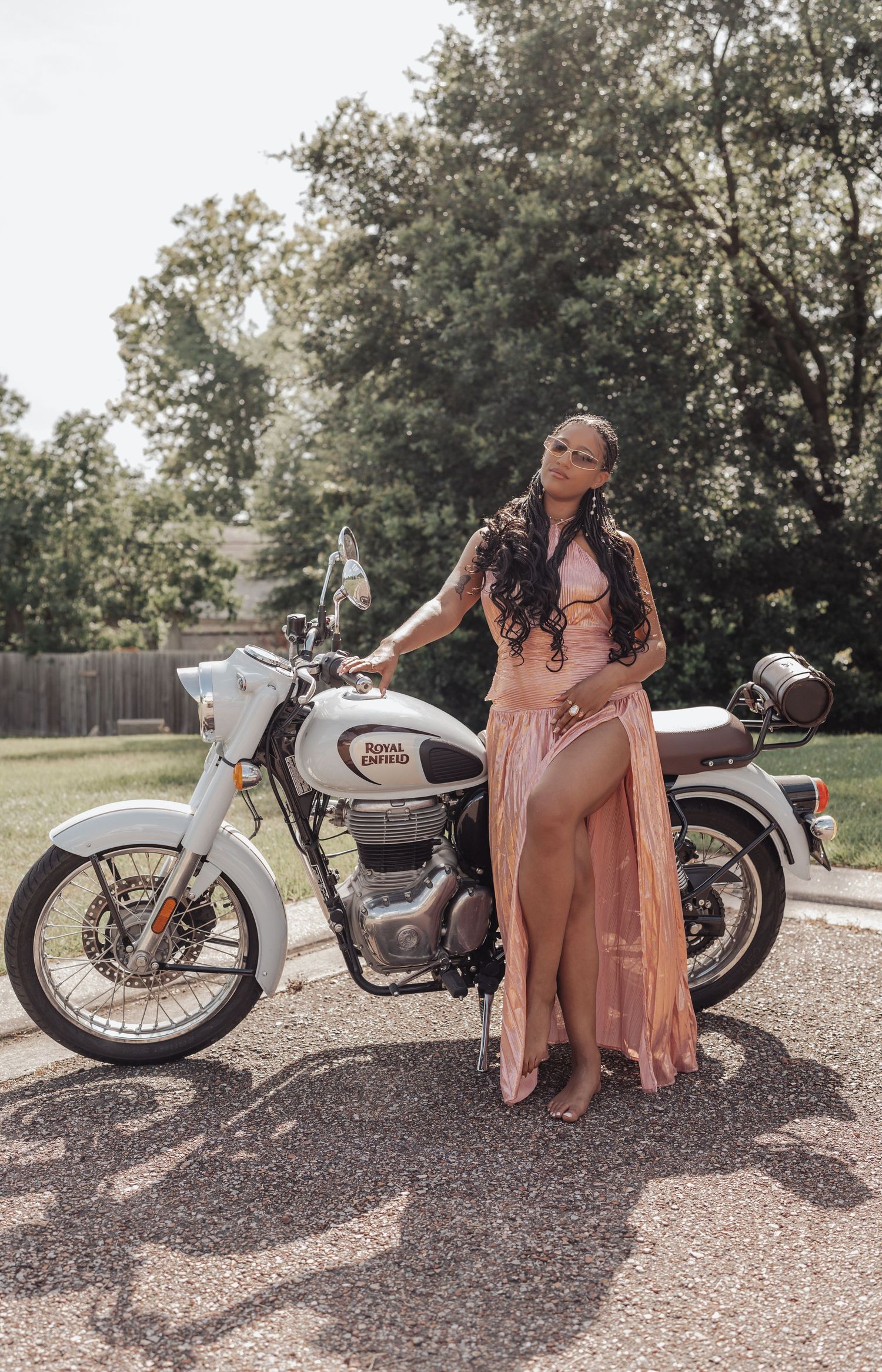 A woman in a pink maxi dress poses beside a white Royal Enfield motorcycle on a paved path surrounded by trees.