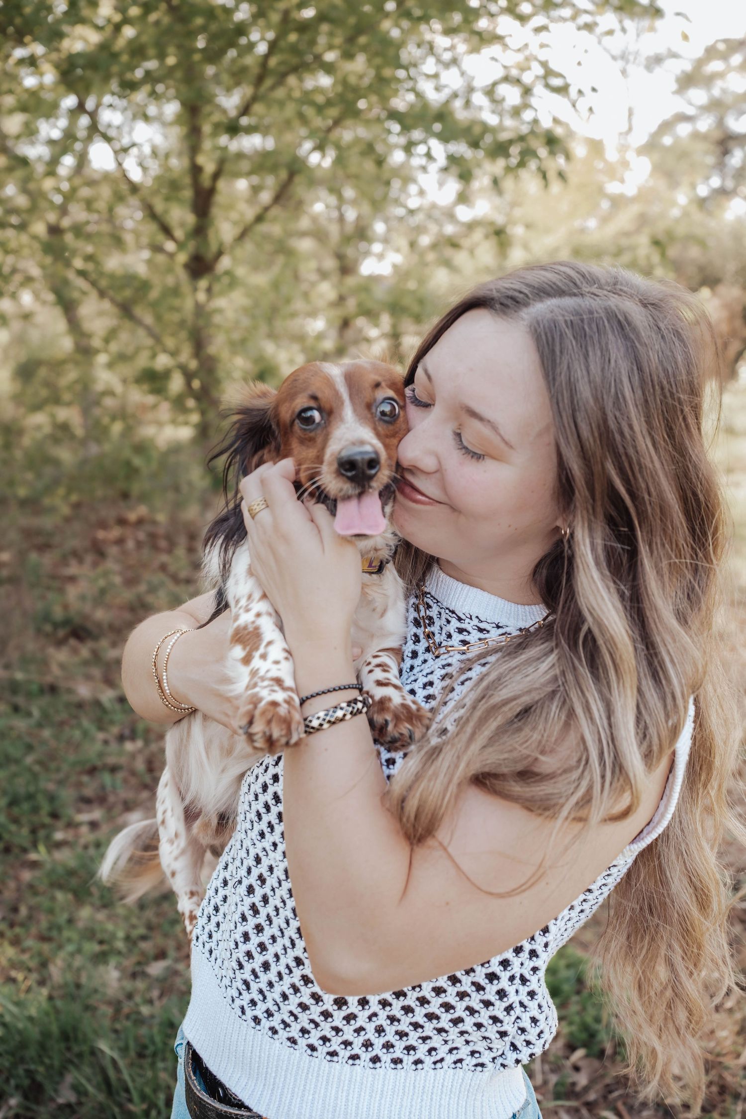 Woman with long hair kissing her brown and white Dachshund puppy outdoors in a forest setting.
