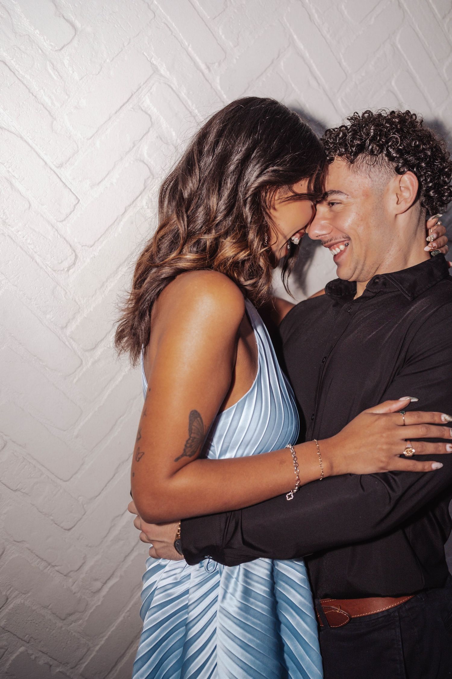 A joyful couple embraces and smiles at each other, woman in blue dress, man in black shirt, against white brick wall.