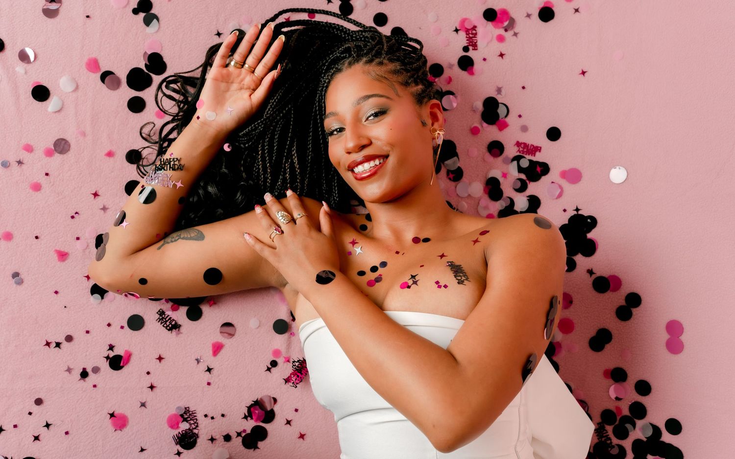 A joyful young woman with braids poses among black and pink confetti against a pink background, wearing a white top.