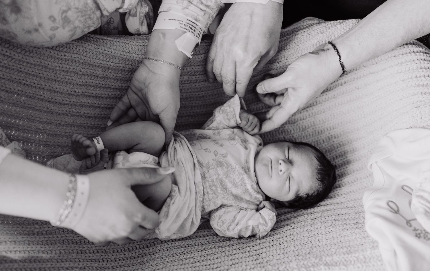 Black and white photo of a newborn baby lying on a knit blanket while several adult hands gently hold and examine the infant.