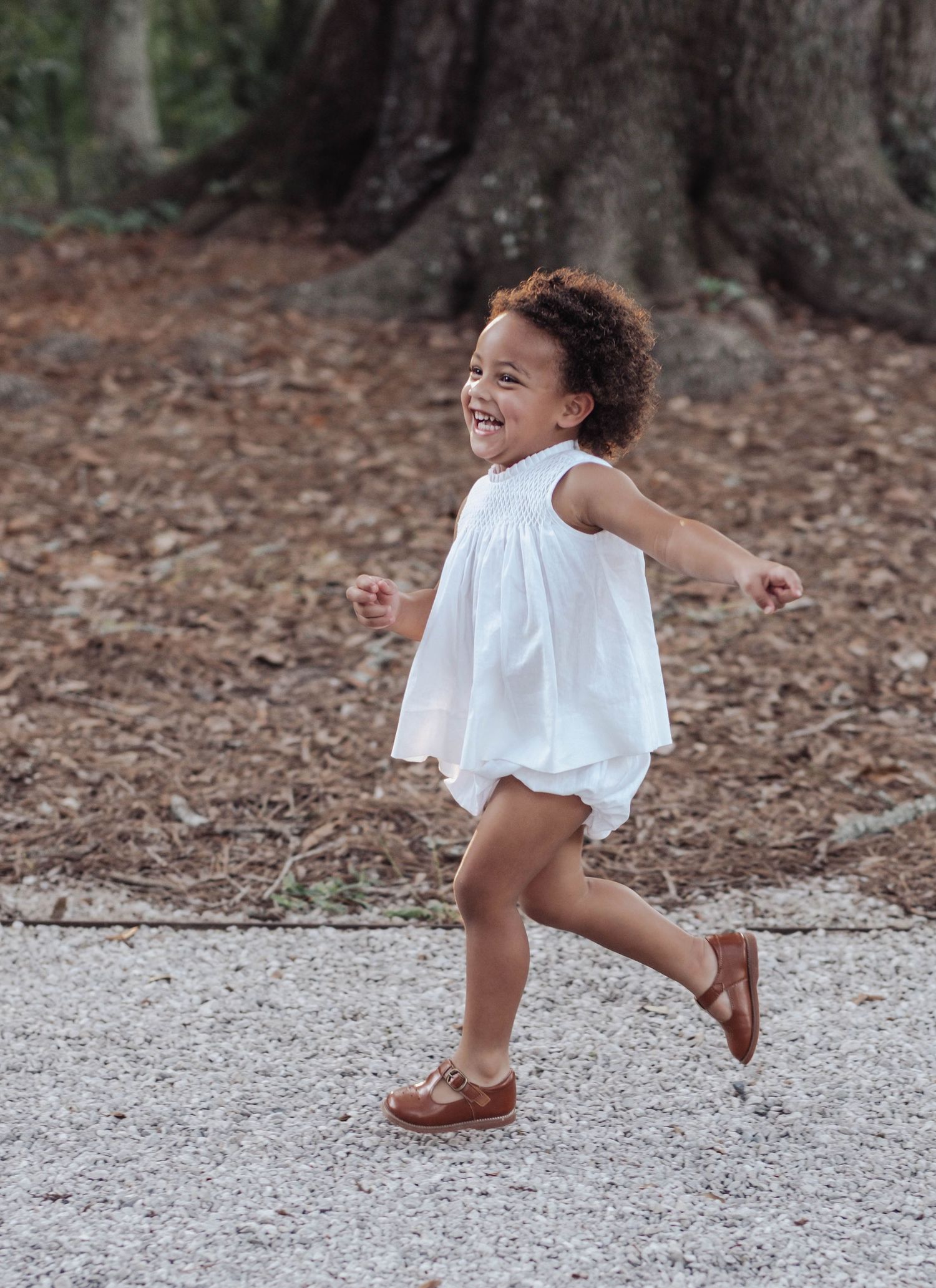 A child in a white dress twirls and dances playfully on a gravel path surrounded by fallen leaves.