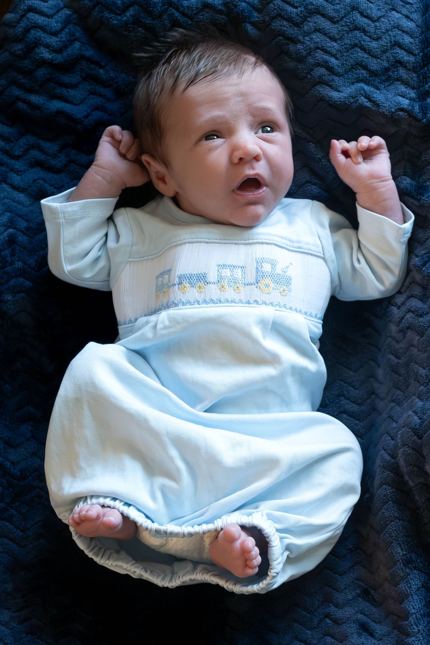 A newborn baby wearing a white romper lies on a dark blue textured blanket in a series of candid poses.