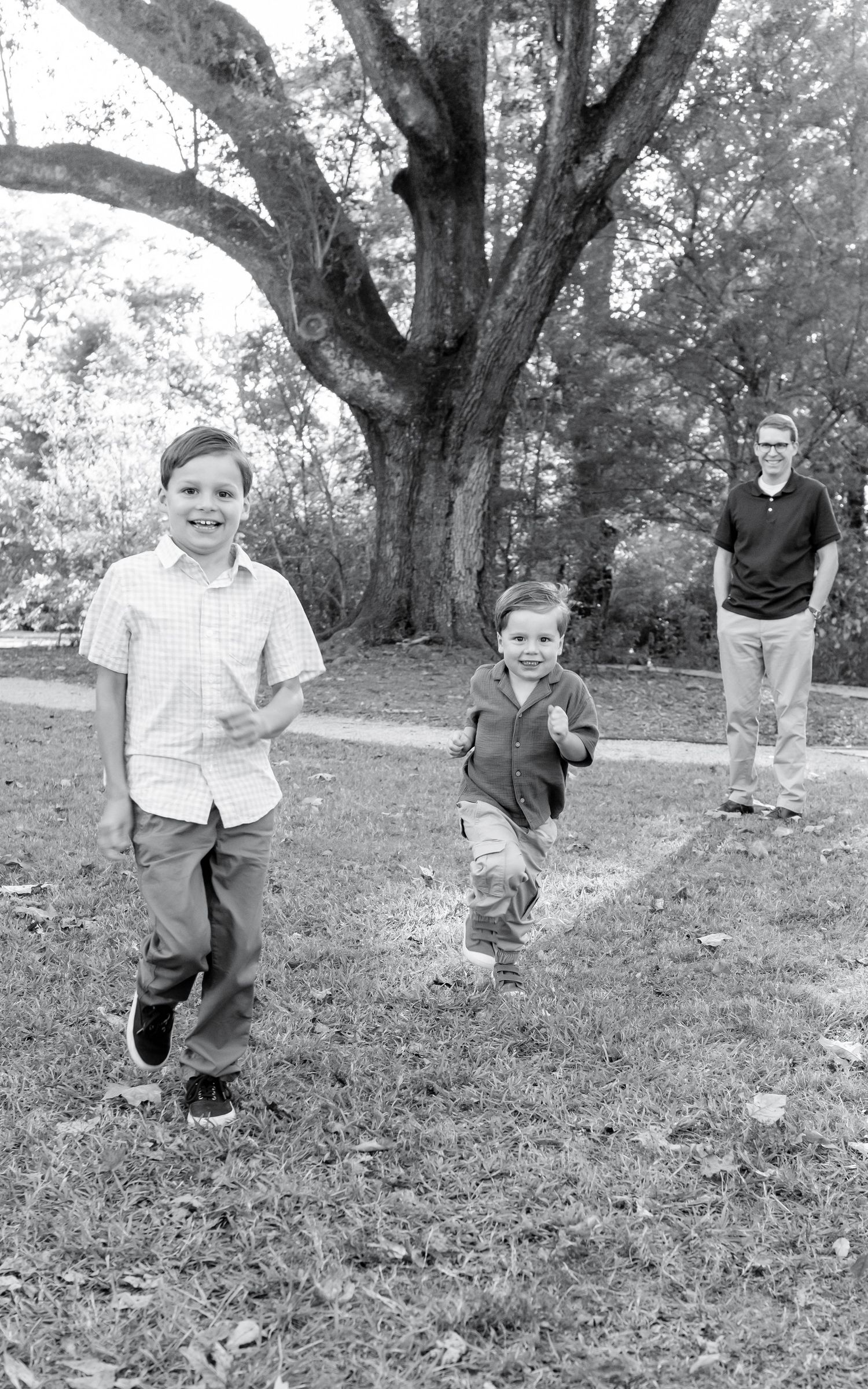 Two boys running toward camera in a park with a large tree, while an older teen watches in background, black and white photo.