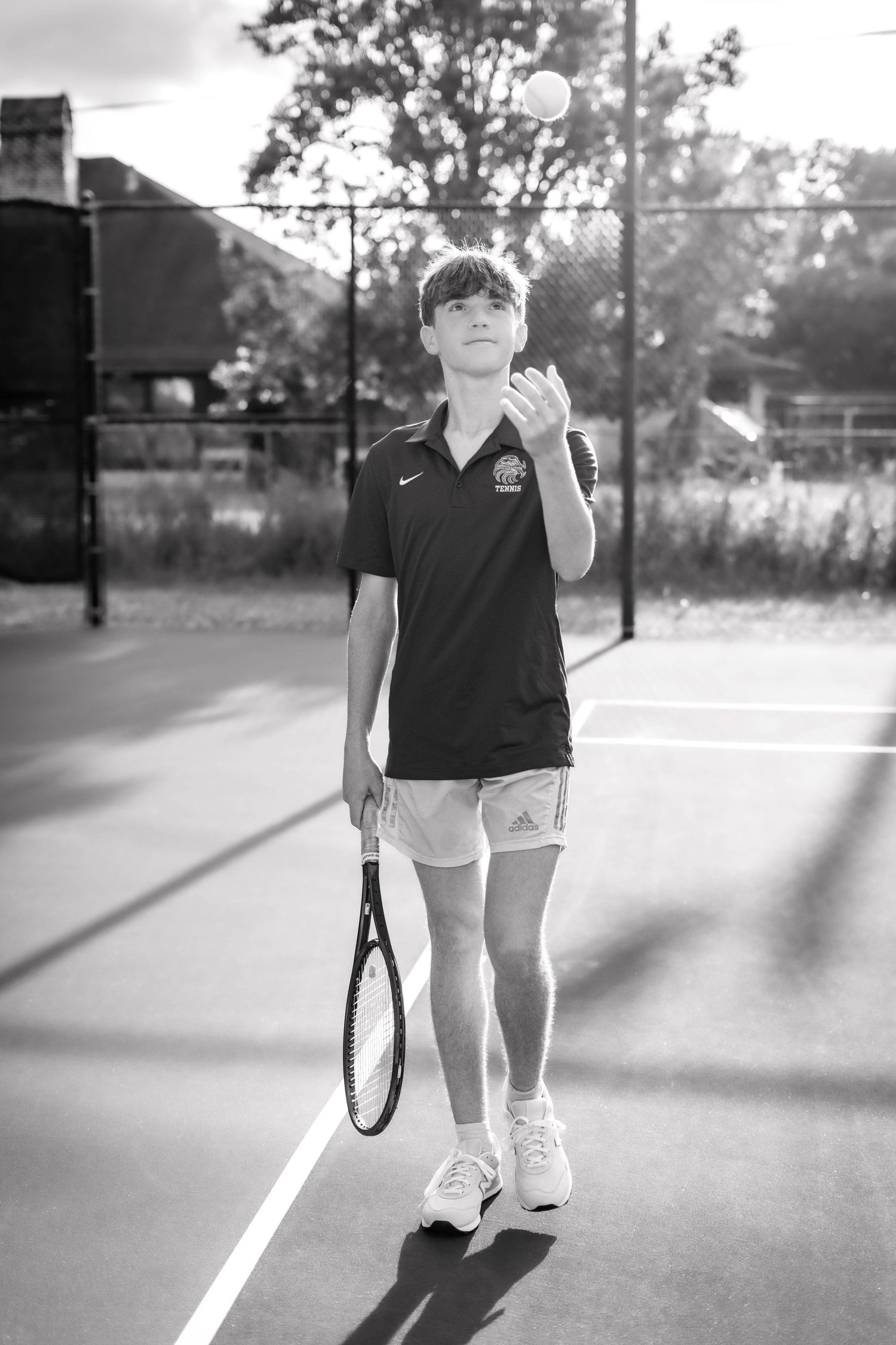 Young tennis player tossing ball to serve on outdoor court, holding racket, wearing Nike shirt and Adidas shorts.