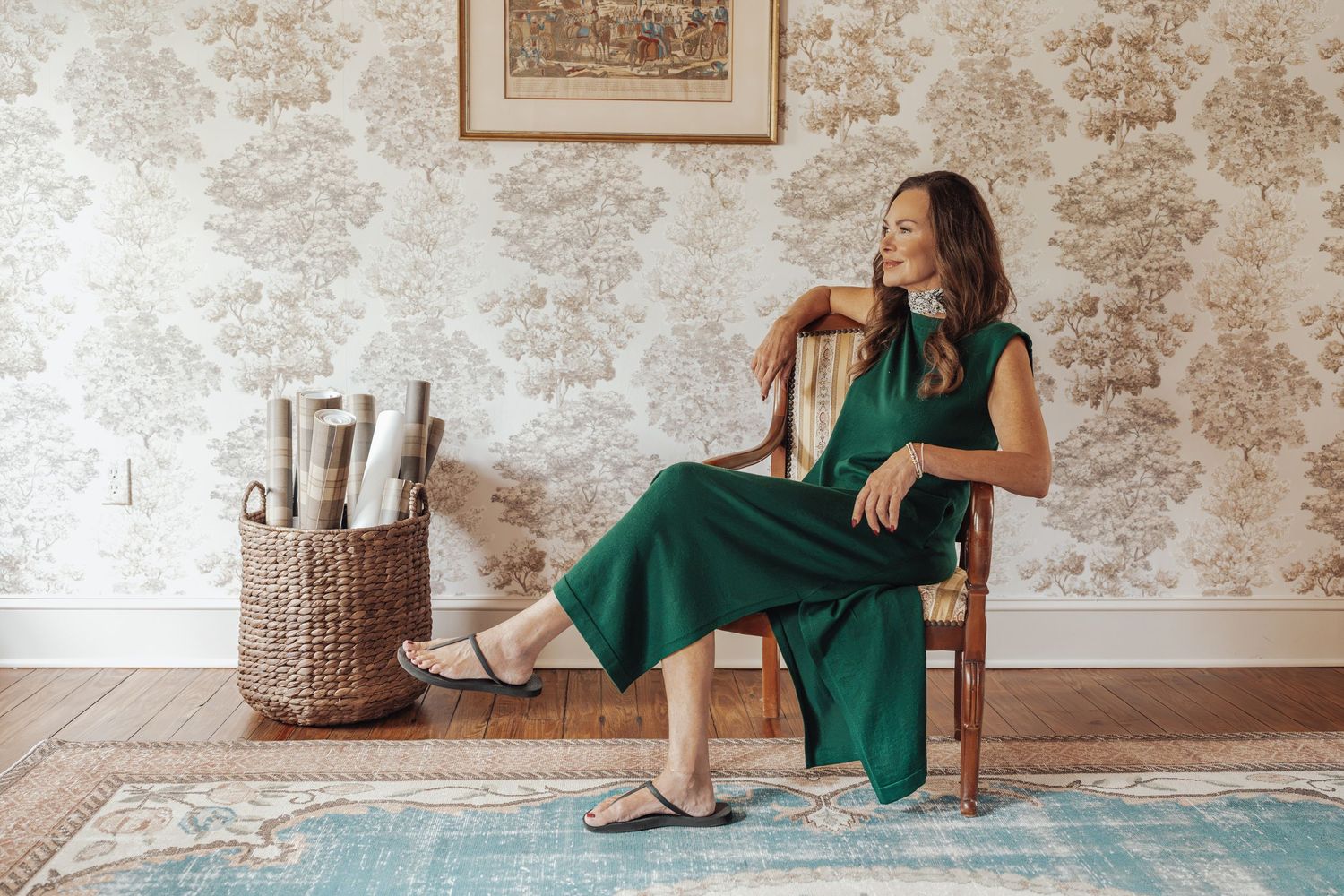 Woman in a green dress sits in a wooden chair by floral toile wallpaper, with a basket of birch logs and a teal vintage rug nearby.