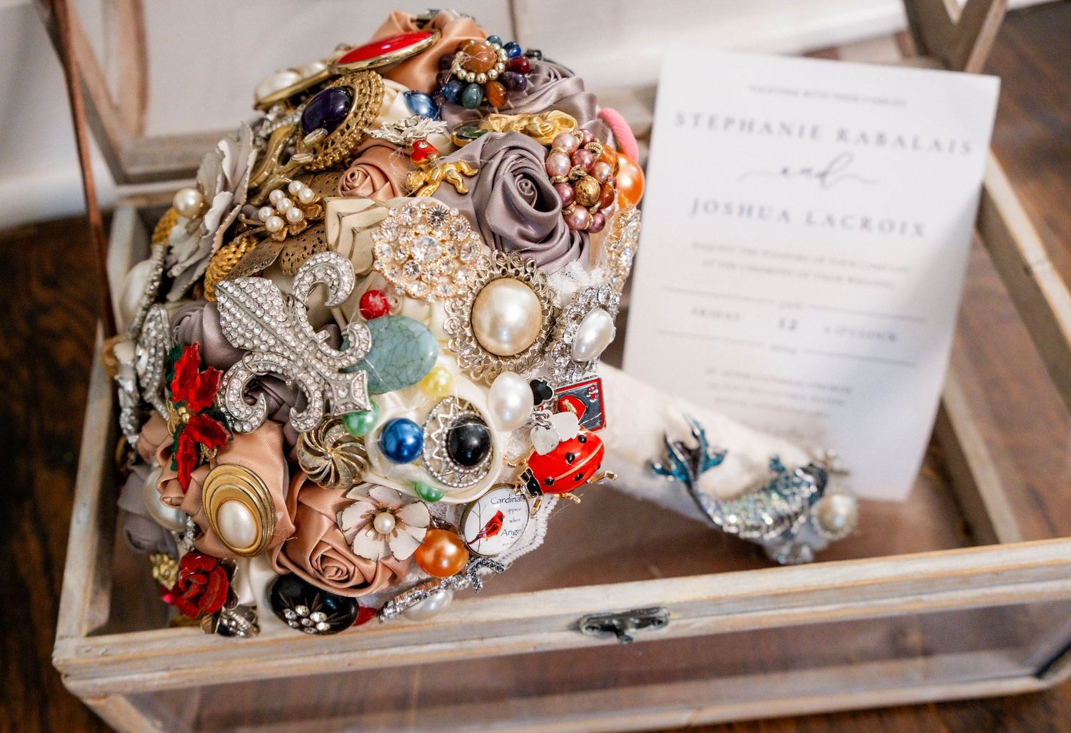 Colorful brooch bouquet with jewels and pearls displayed in a glass box beside a wedding invitation.