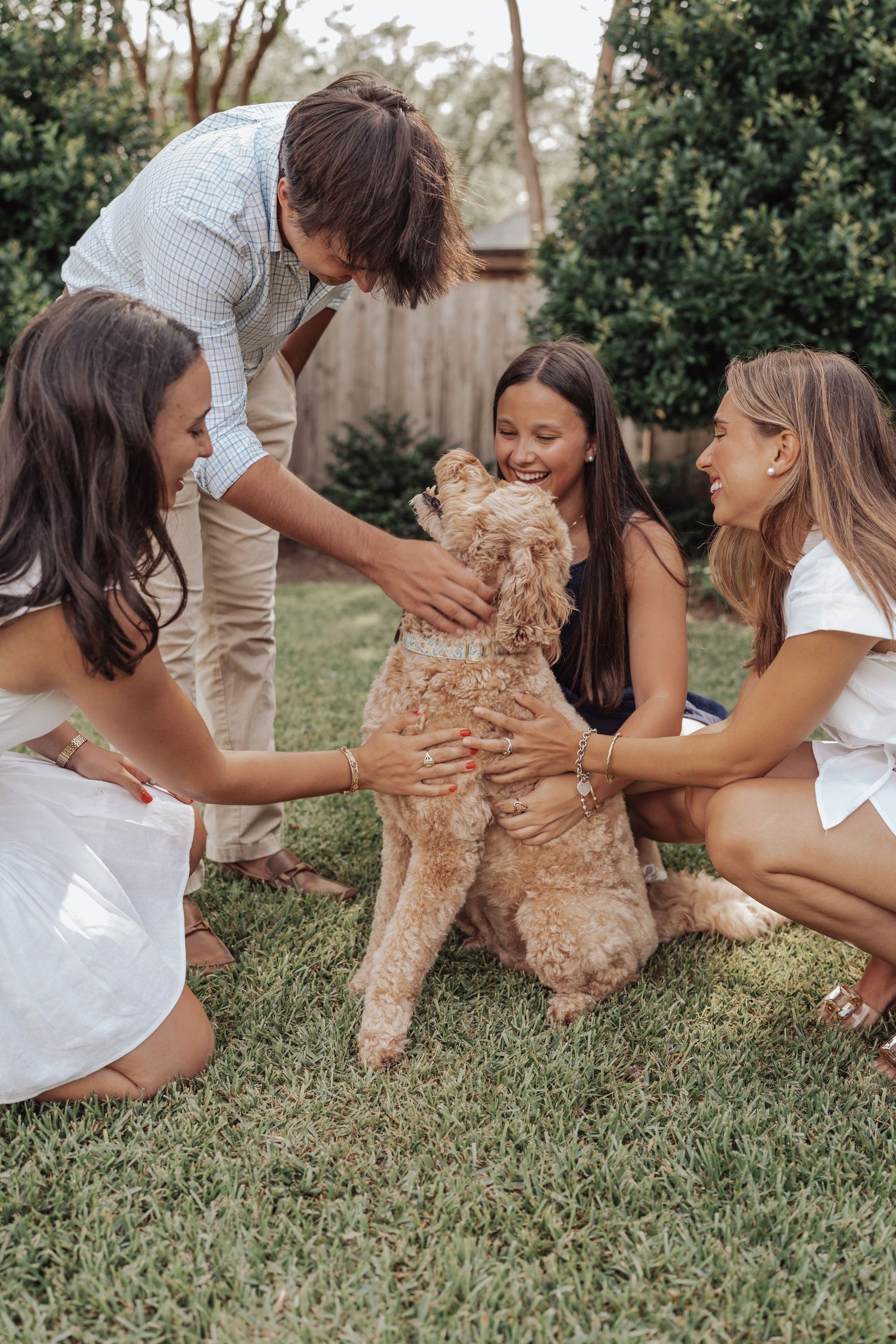 Family of four petting their fluffy golden doodle dog together on green grass in a backyard garden.