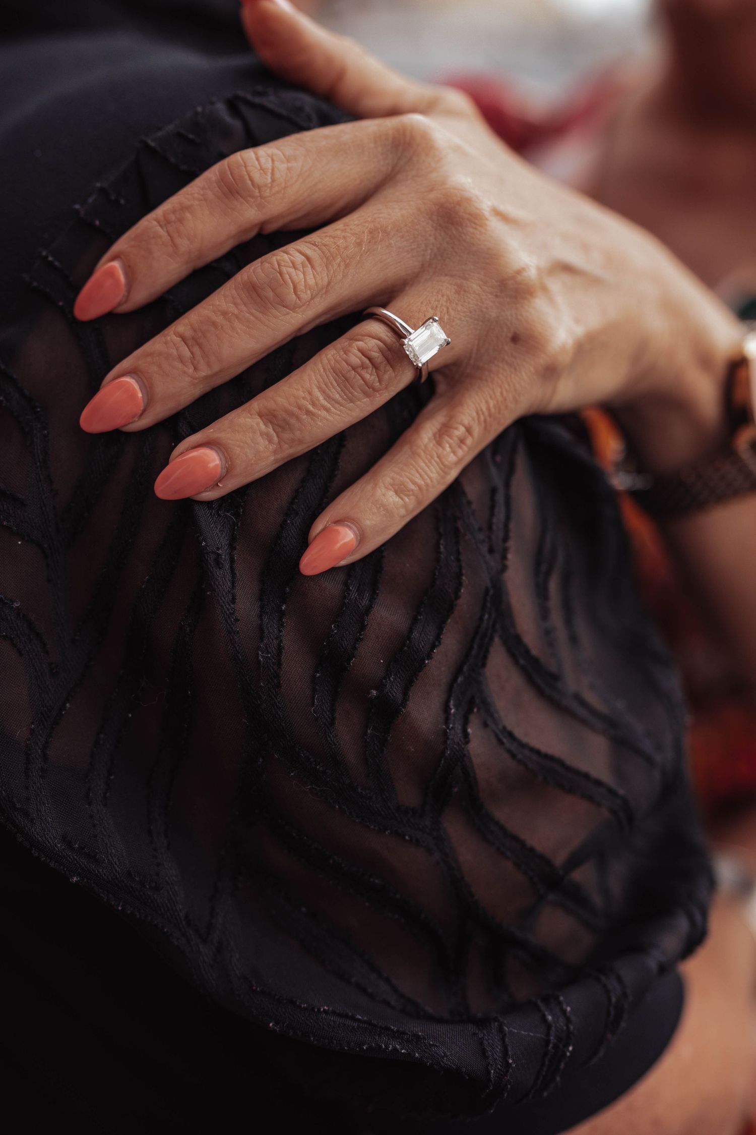 Close-up of a hand with coral nails wearing a square diamond ring, resting on black sheer fabric.