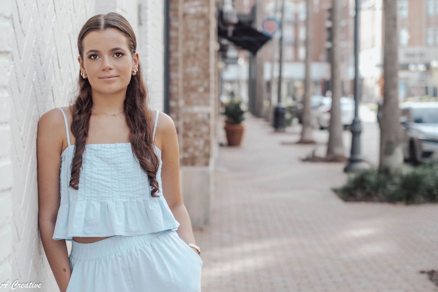 A person wearing a light blue summer dress stands on a city sidewalk near brick buildings and parked cars.