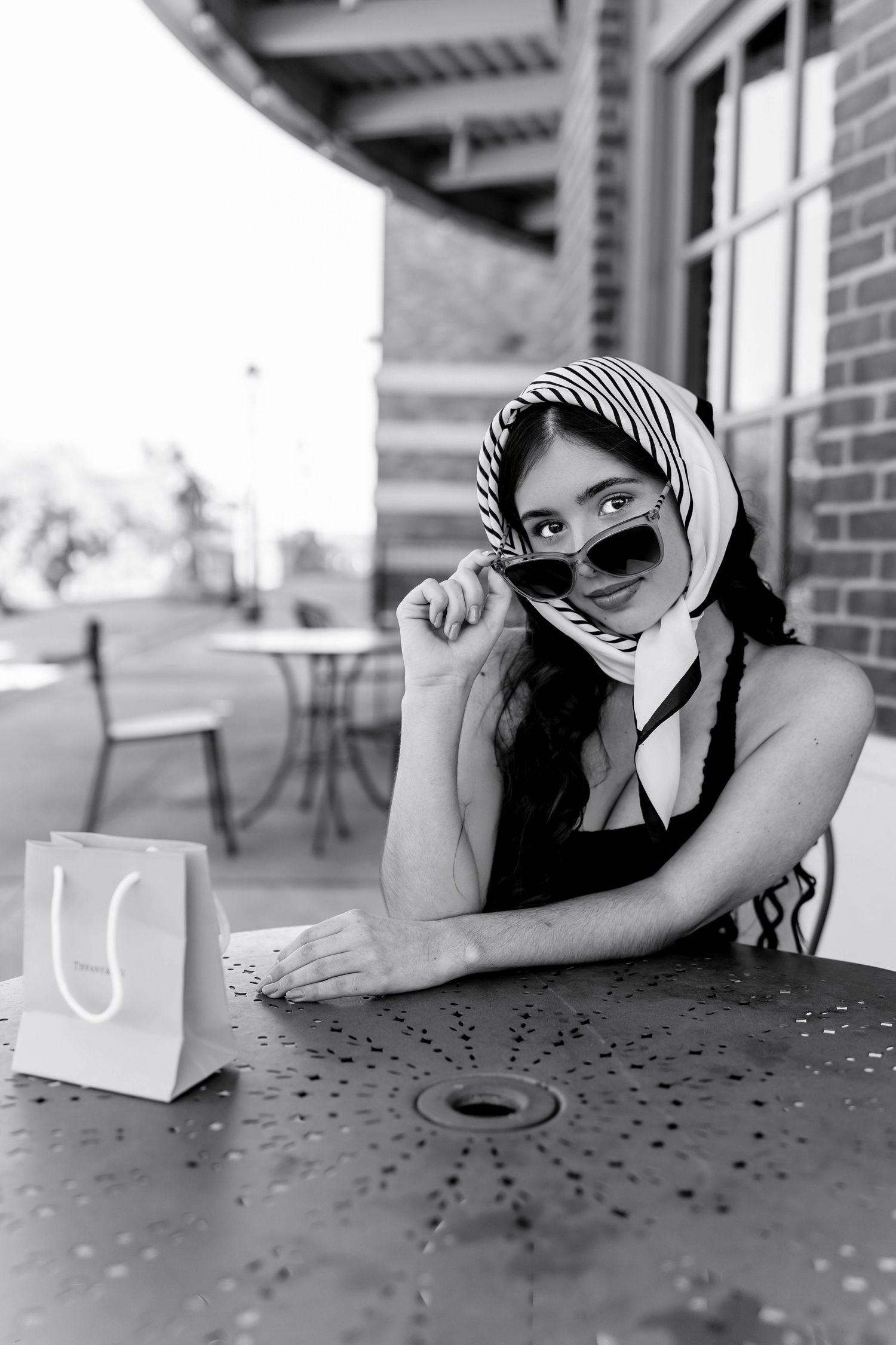Black and white photo of a stylish woman in sunglasses and headscarf sitting at an outdoor cafe table.