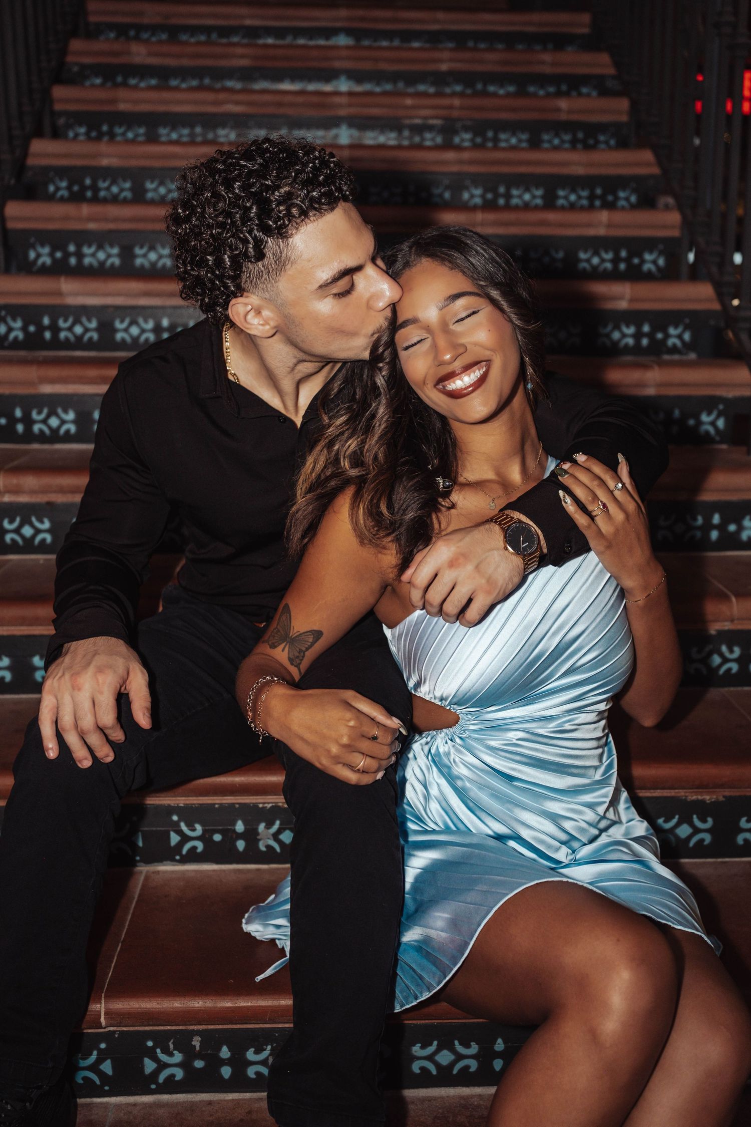 A man in black kisses a smiling woman in a blue satin dress on the cheek while sitting on decorative stairs.