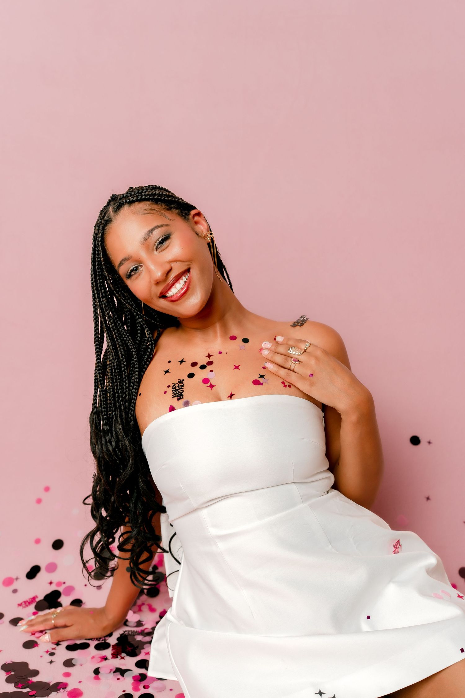 Smiling woman with long braids in white strapless dress, covered in colorful confetti against pink background.