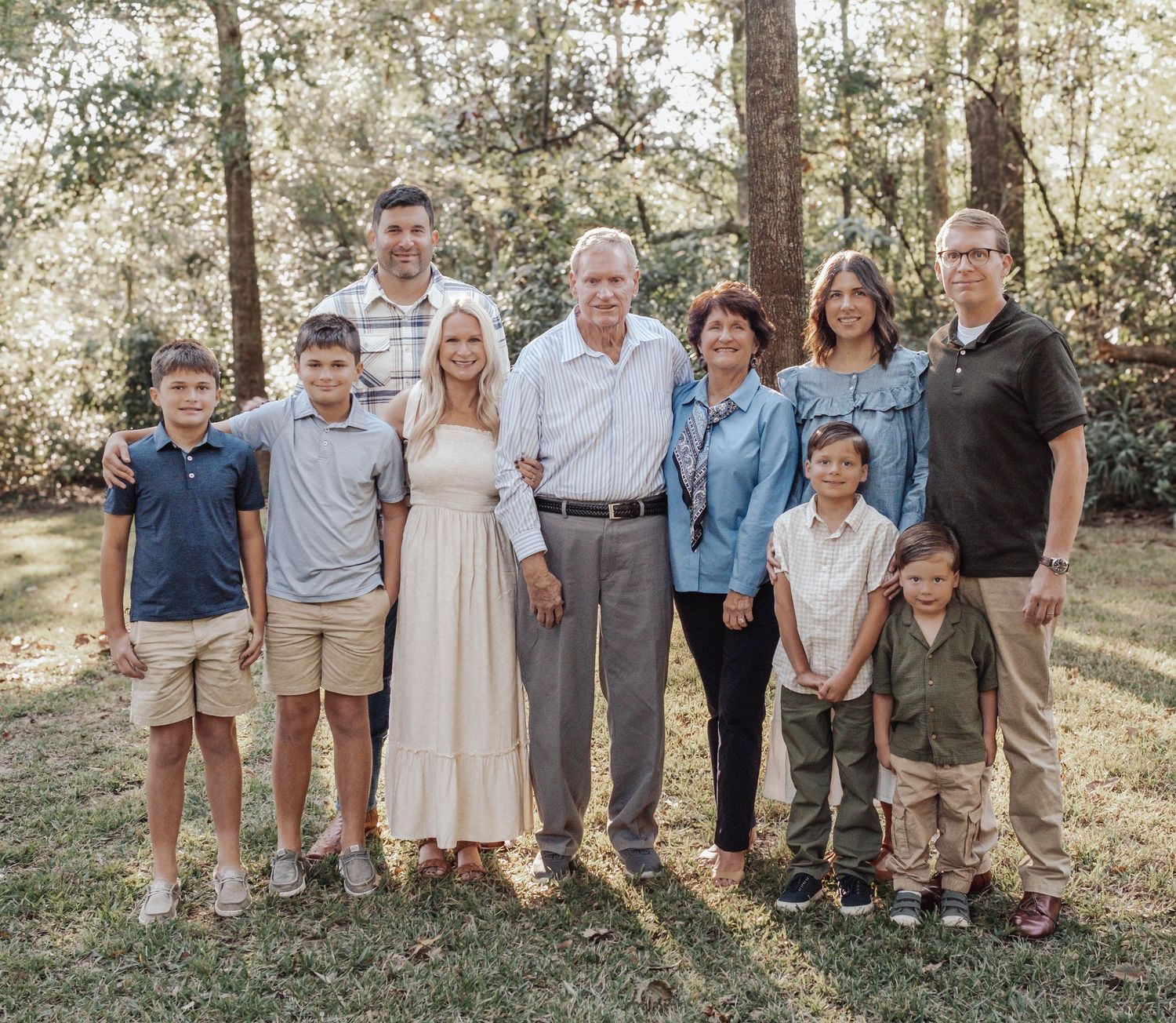 Multi-generational family portrait outdoors, smiling group of adults and children posing together in a wooded backyard setting.