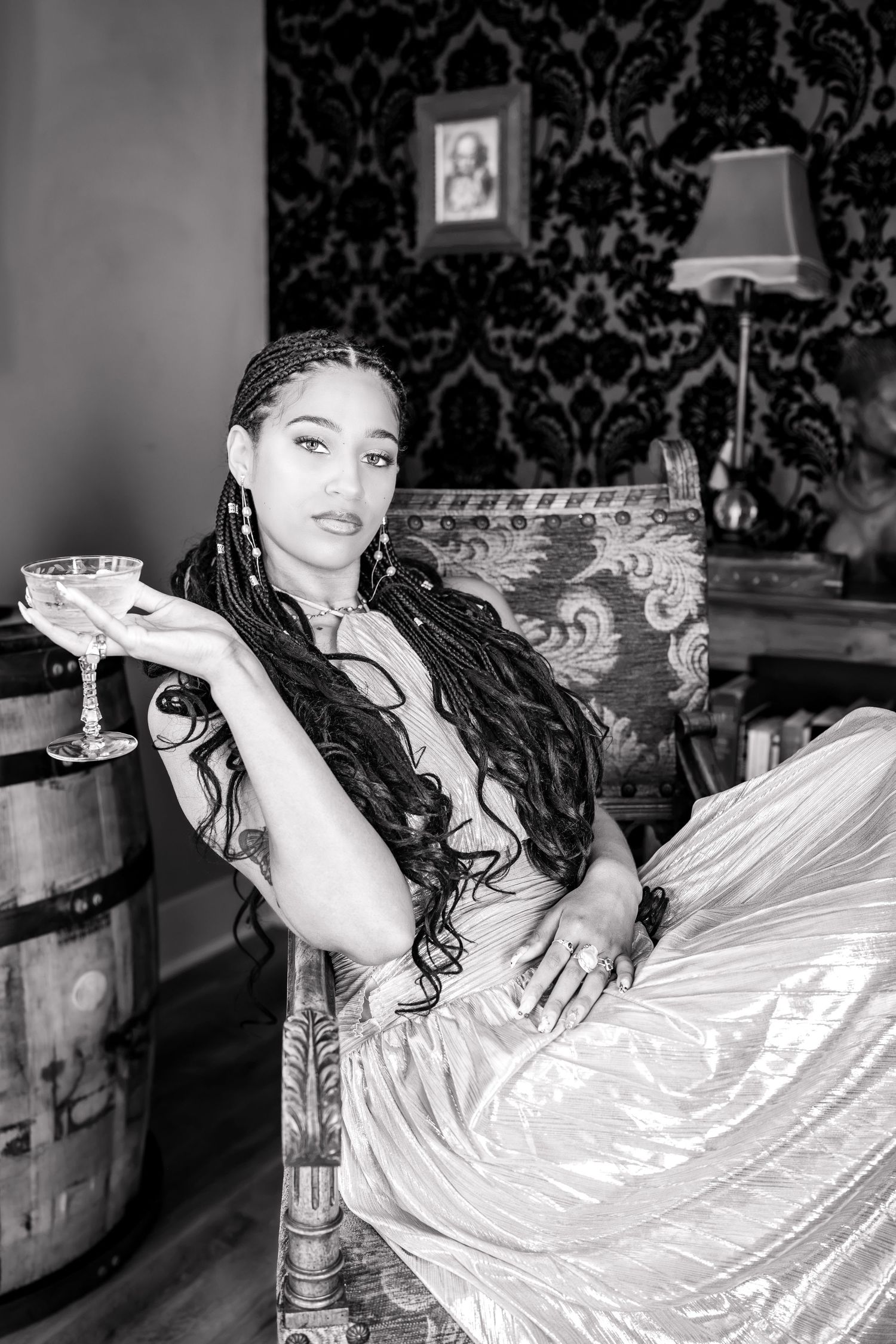 Black and white portrait of a woman with long braids holding a cocktail glass, wearing a flowing dress in an elegant room.