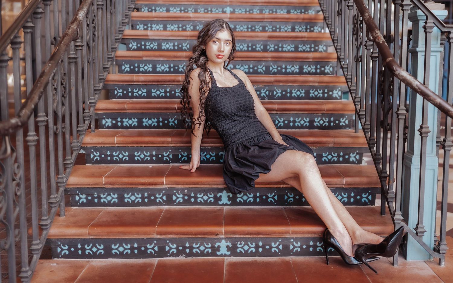 A young woman in a black dress and heels poses gracefully on decorative tiled stairs with ornate railings.