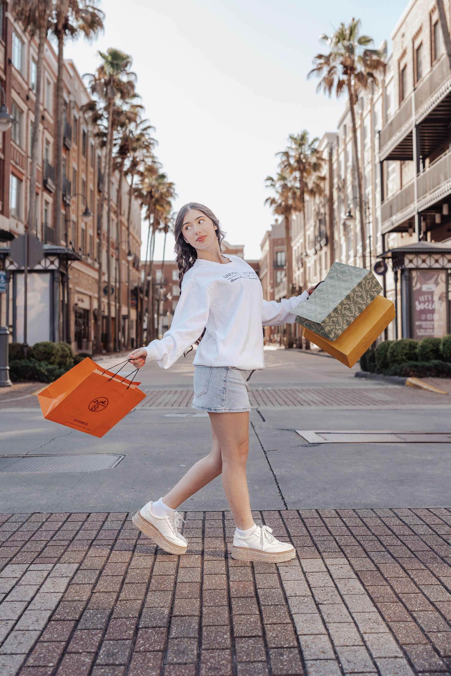 Young woman in white hoodie and denim shorts carrying colorful shopping bags on a palm-lined outdoor shopping street.