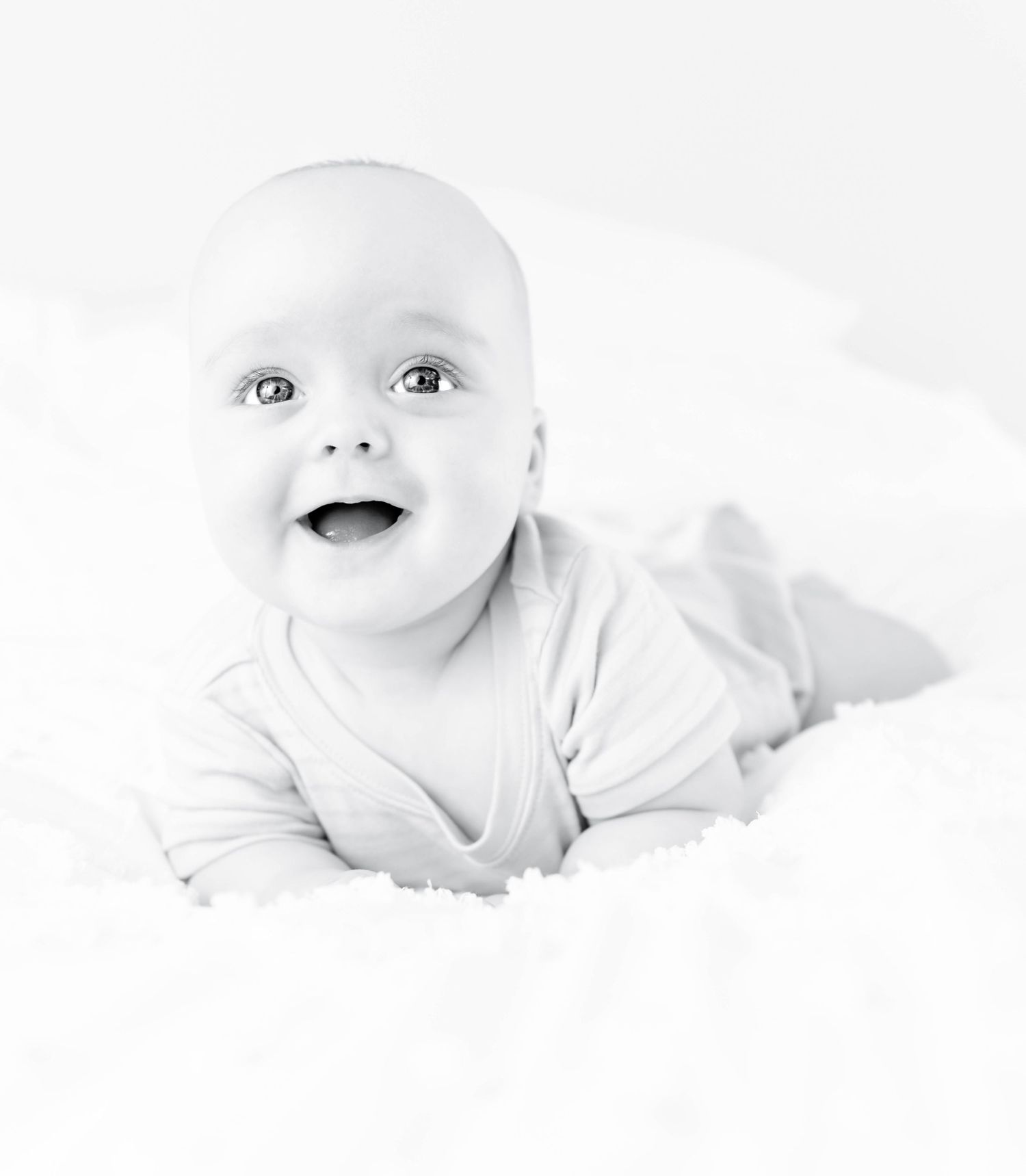 Adorable baby in white clothing lies on white backdrop smiling in black and white portrait.