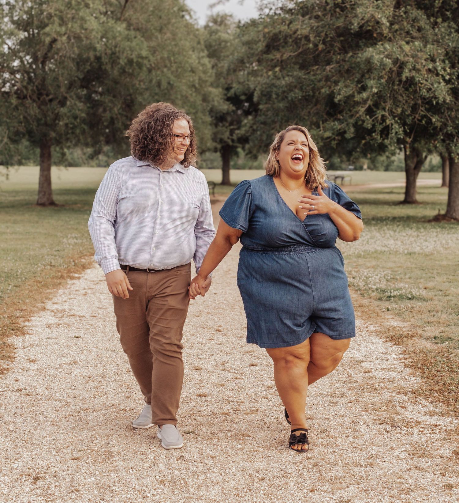 Happy couple walking together and laughing on a gravel path in a park surrounded by trees.