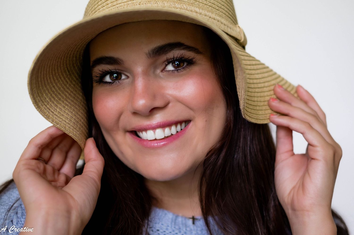 A smiling person in a straw sun hat and denim shirt touches the brim of their hat against a white background.
