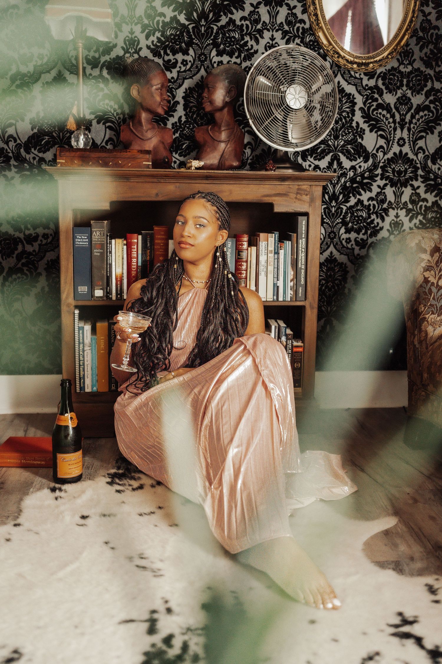 Woman in pink satin dress holding wine glass, seated by bookshelf with decor, fan, and patterned wallpaper.