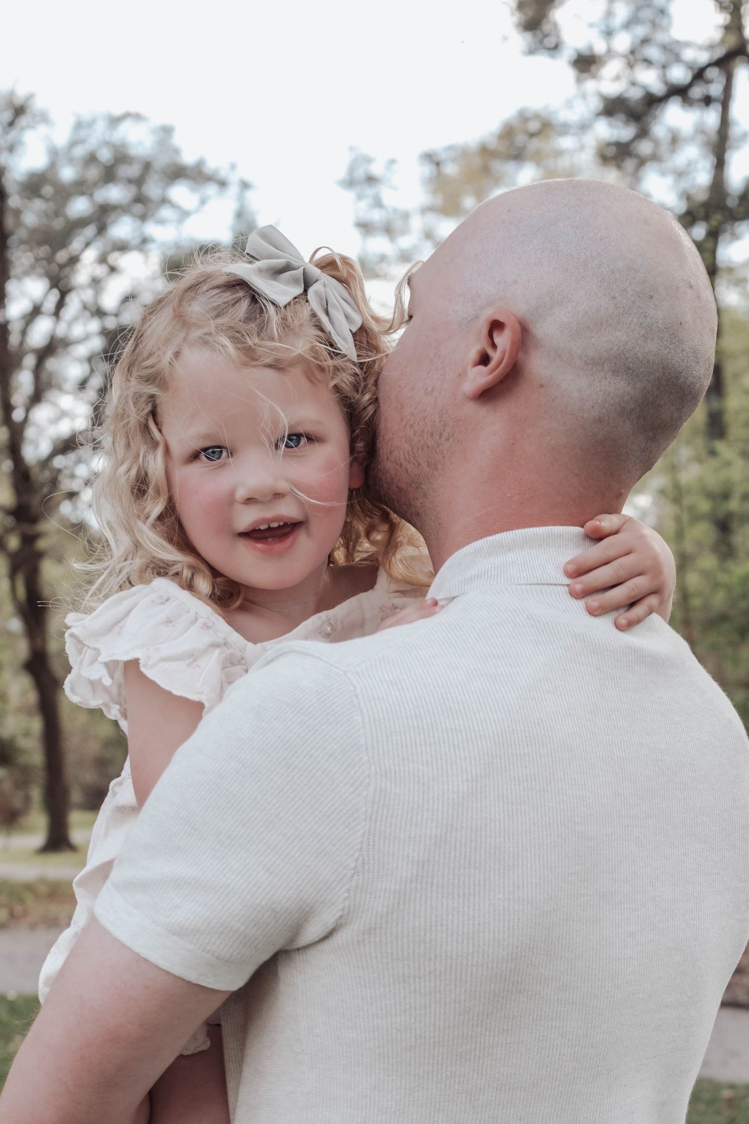 A bald father kisses his curly-haired blonde toddler daughter wearing a pink dress and bow outdoors.
