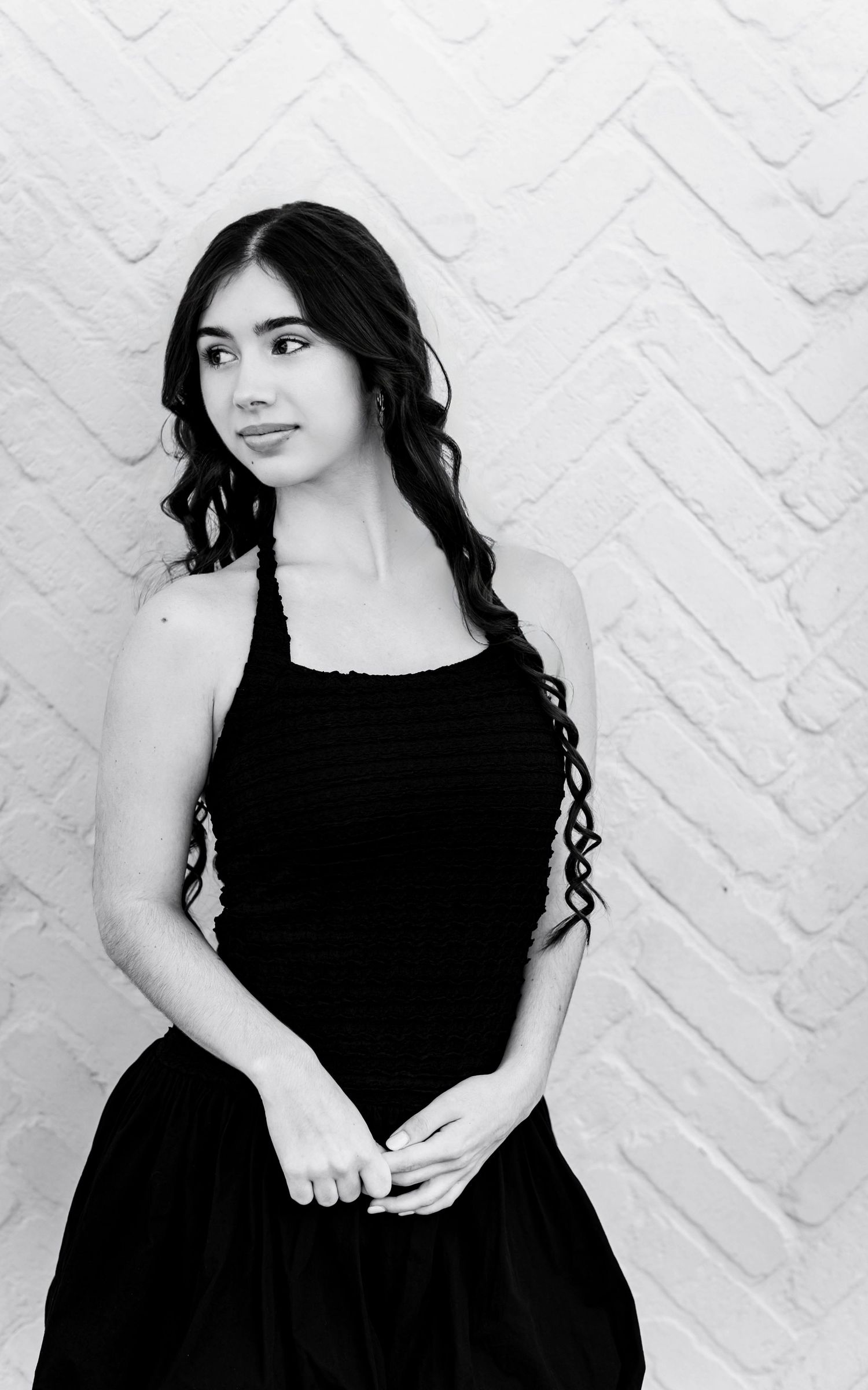 Black and white portrait of a young woman in a black halter dress, posing gracefully against a white brick wall.