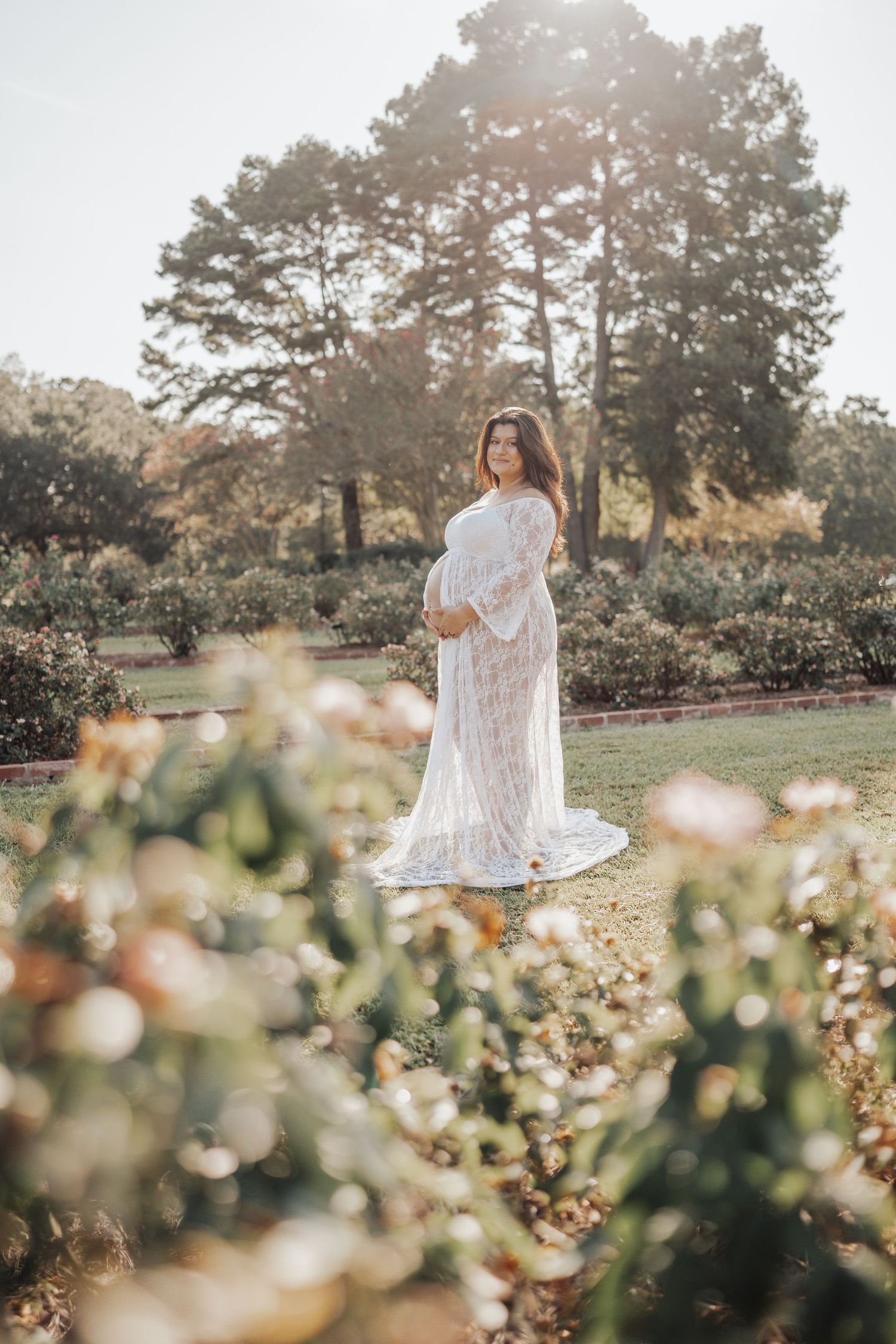 Pregnant woman in white lace gown standing in sunlit garden with blooming flowers and trees.