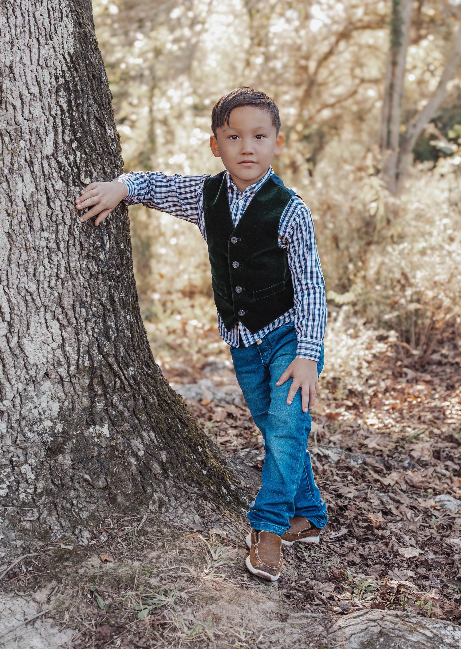 A young child in a vest and plaid shirt leans against a large tree trunk in an autumn woodland setting.
