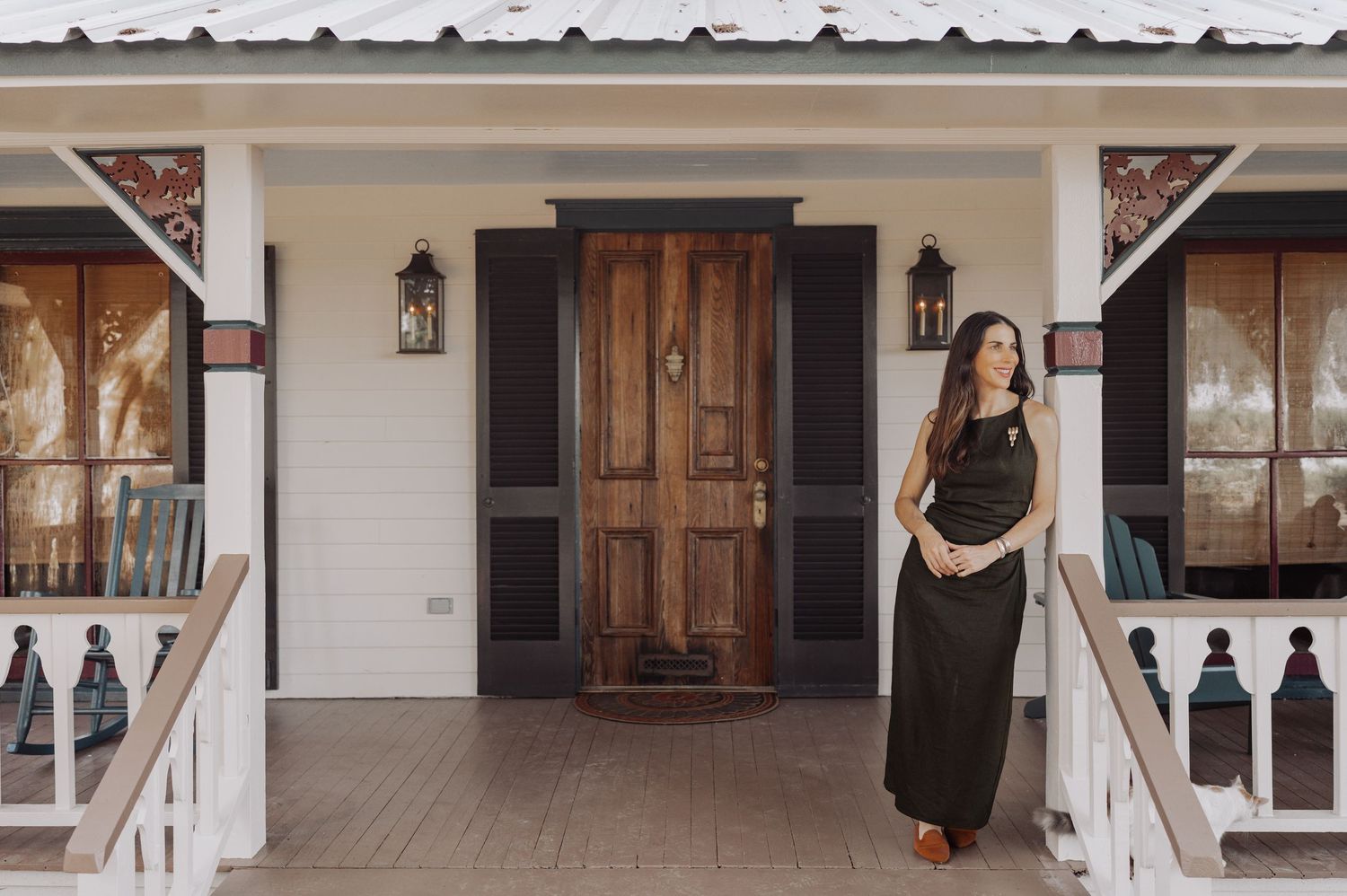 Woman in a long dark dress stands on the wooden porch of a white farmhouse with a rustic wood front door and lantern sconces.