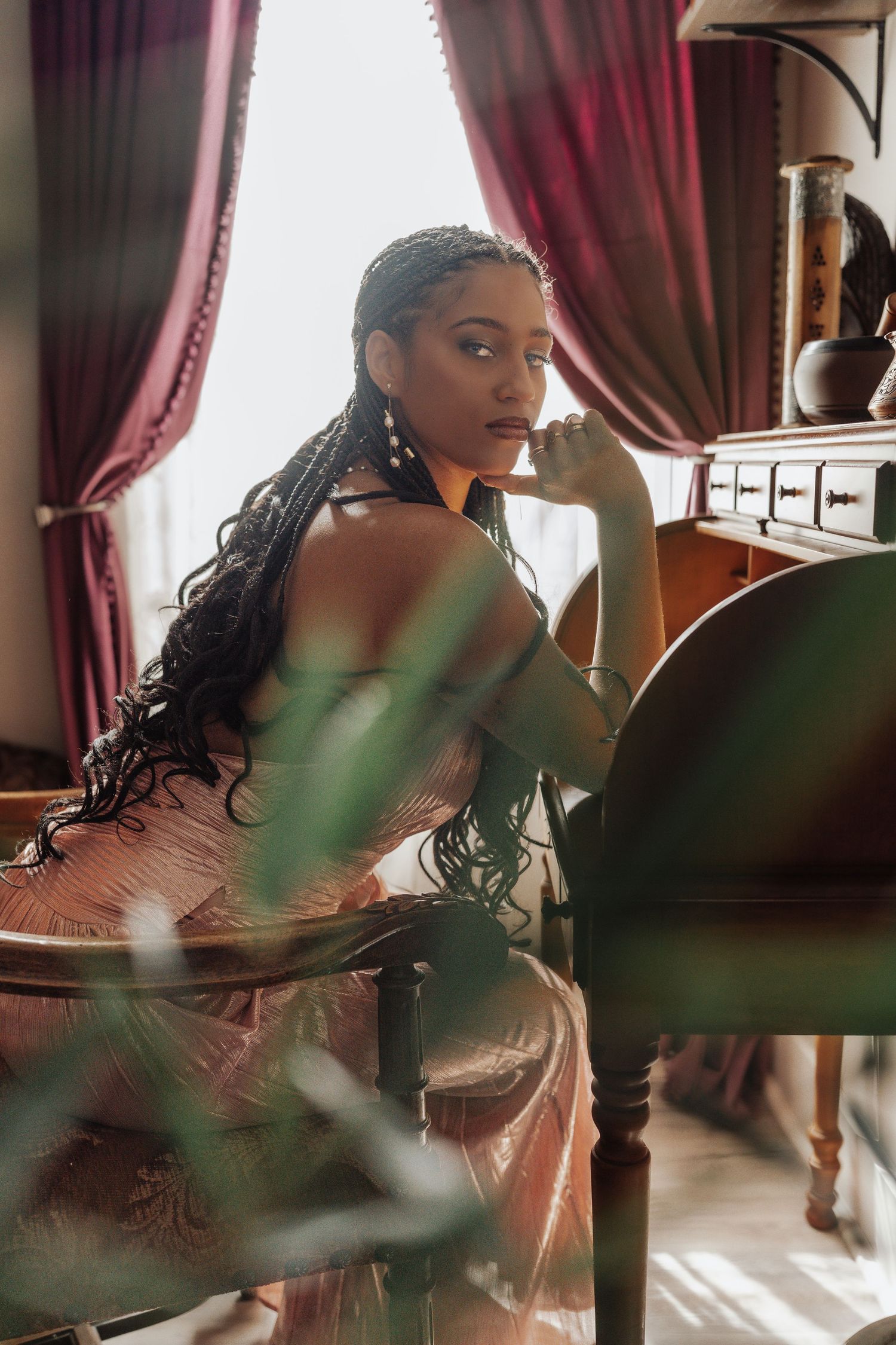 A woman with long braids in a pink dress sits elegantly at a vintage table near red curtains in a warmly lit room.