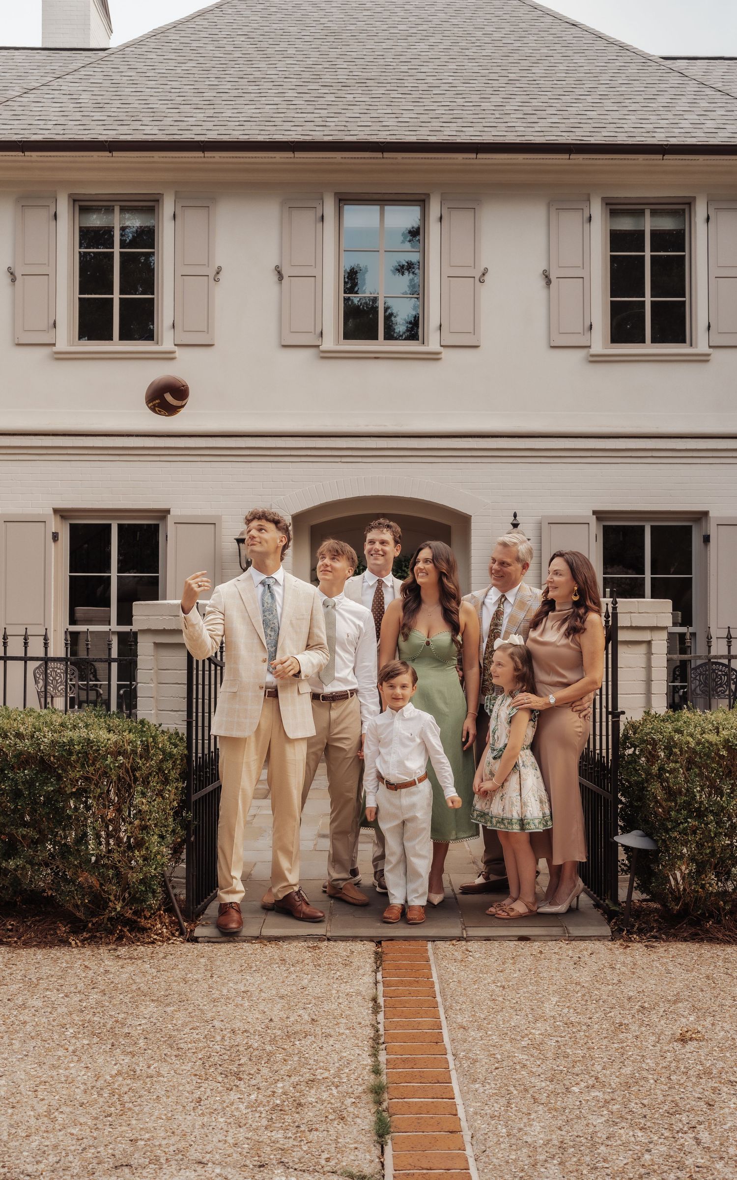 A joyful family group poses outside an elegant beige home, dressed in formal attire for a special occasion.