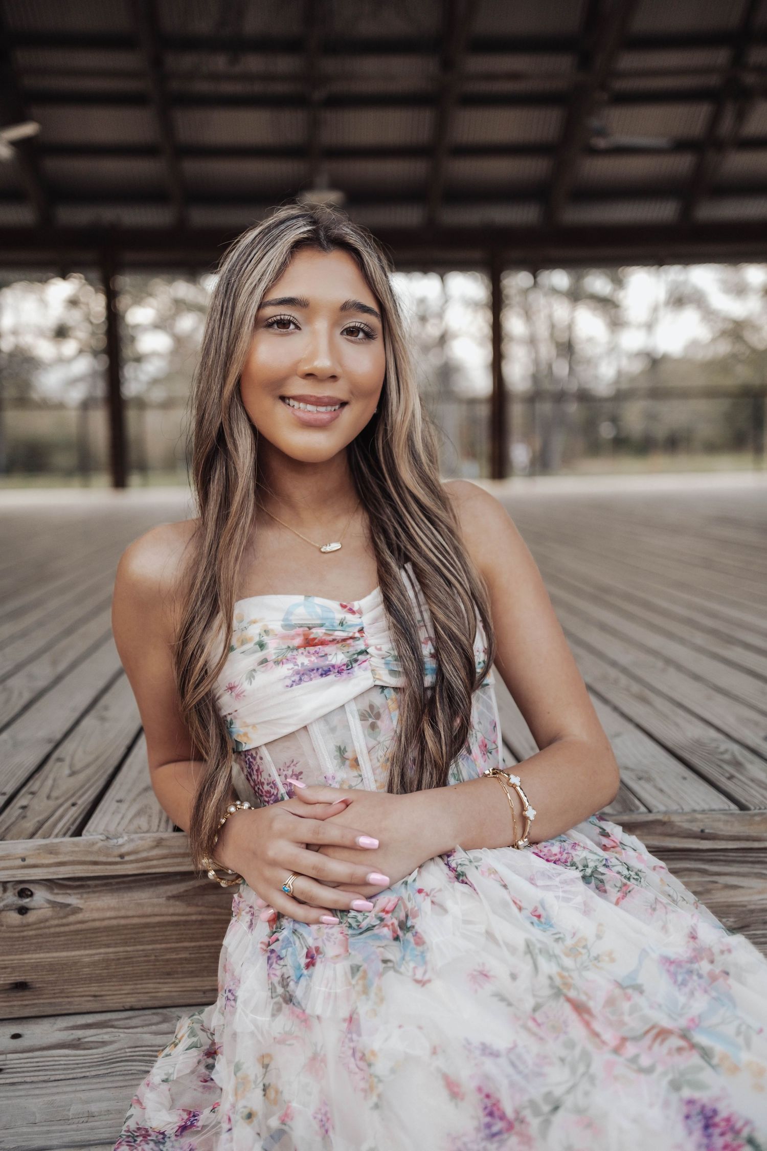 Young woman in floral strapless dress sitting on wooden deck, smiling, with long highlighted hair and gold jewelry.