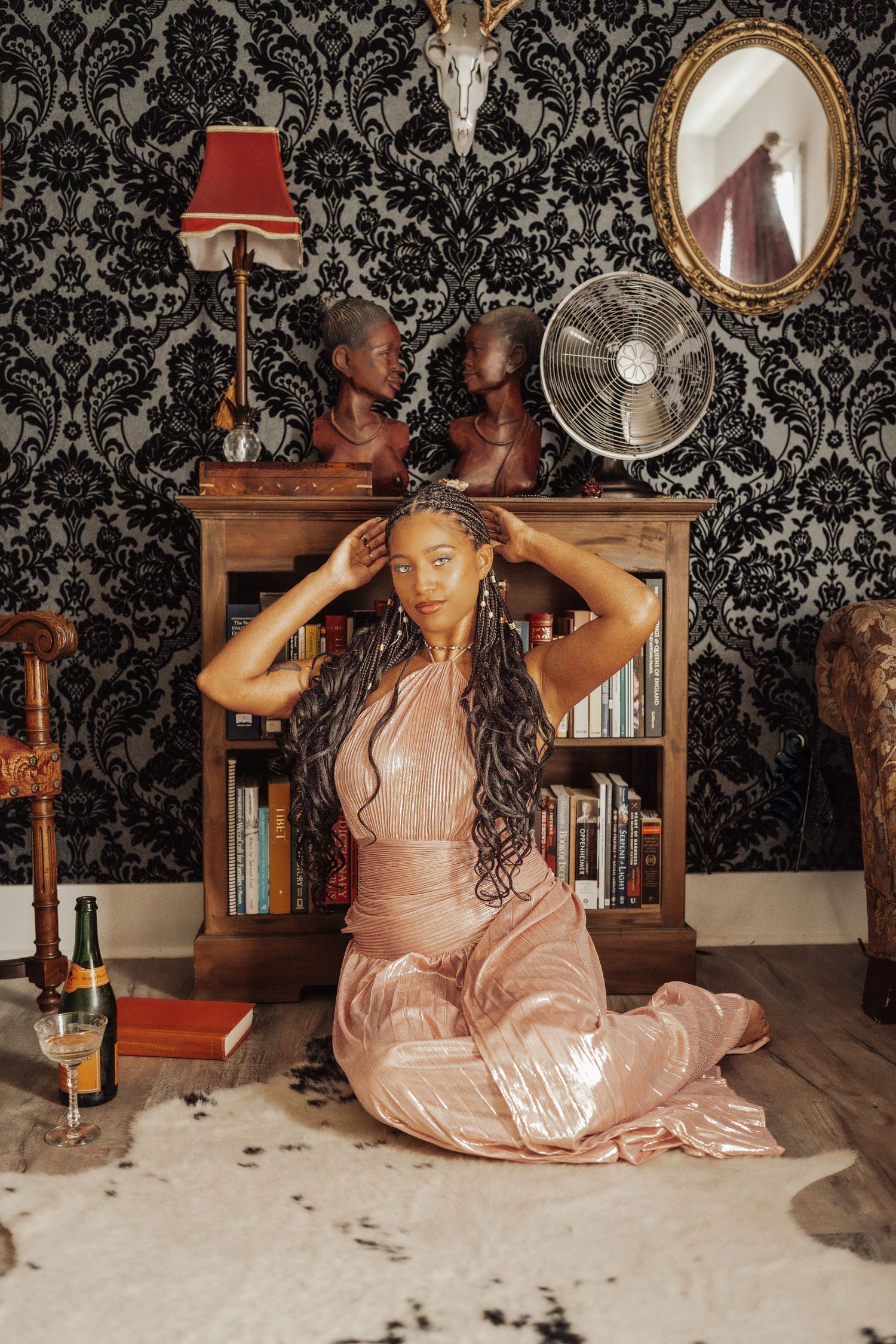 A woman in a pink satin dress is sitting on a fur rug in an eclectic room with a bookcase, mirror, and decorative busts.