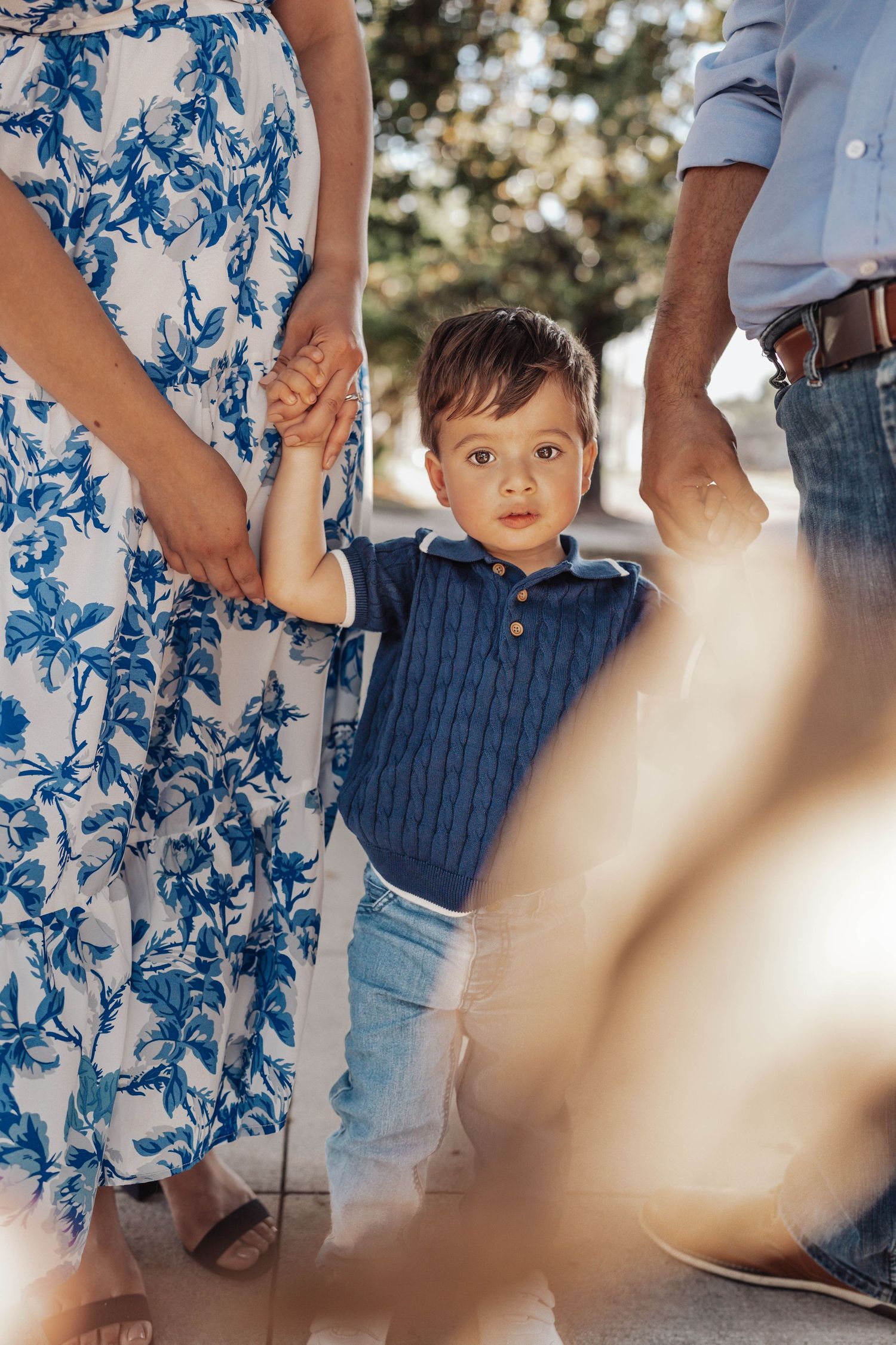 A young toddler boy in a blue knit shirt holds hands with his parents outdoors in warm sunlight.