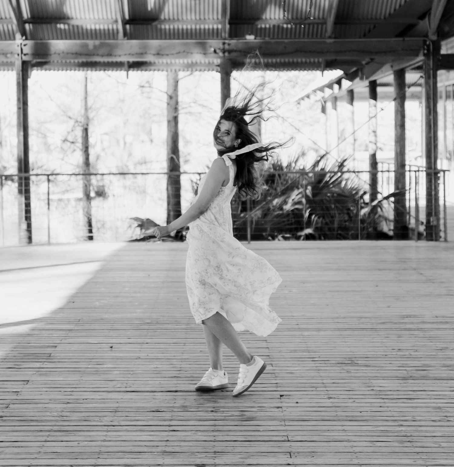 Black and white photo of a young woman in a white dress and sneakers spinning on a wooden deck outdoors.