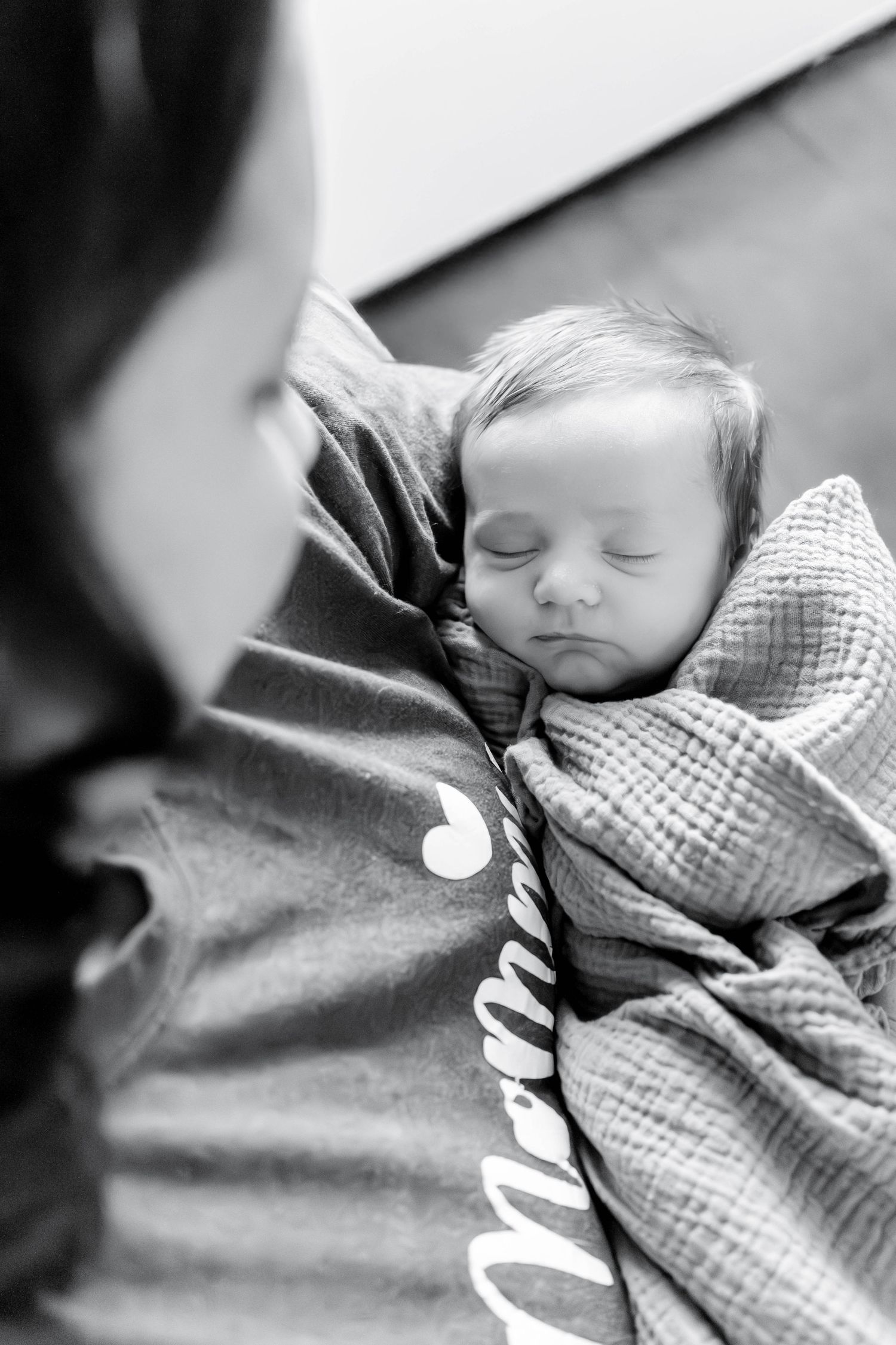 Black and white photo of a sleeping newborn baby wrapped in a blanket, cradled in a mother's arms wearing a mama shirt.