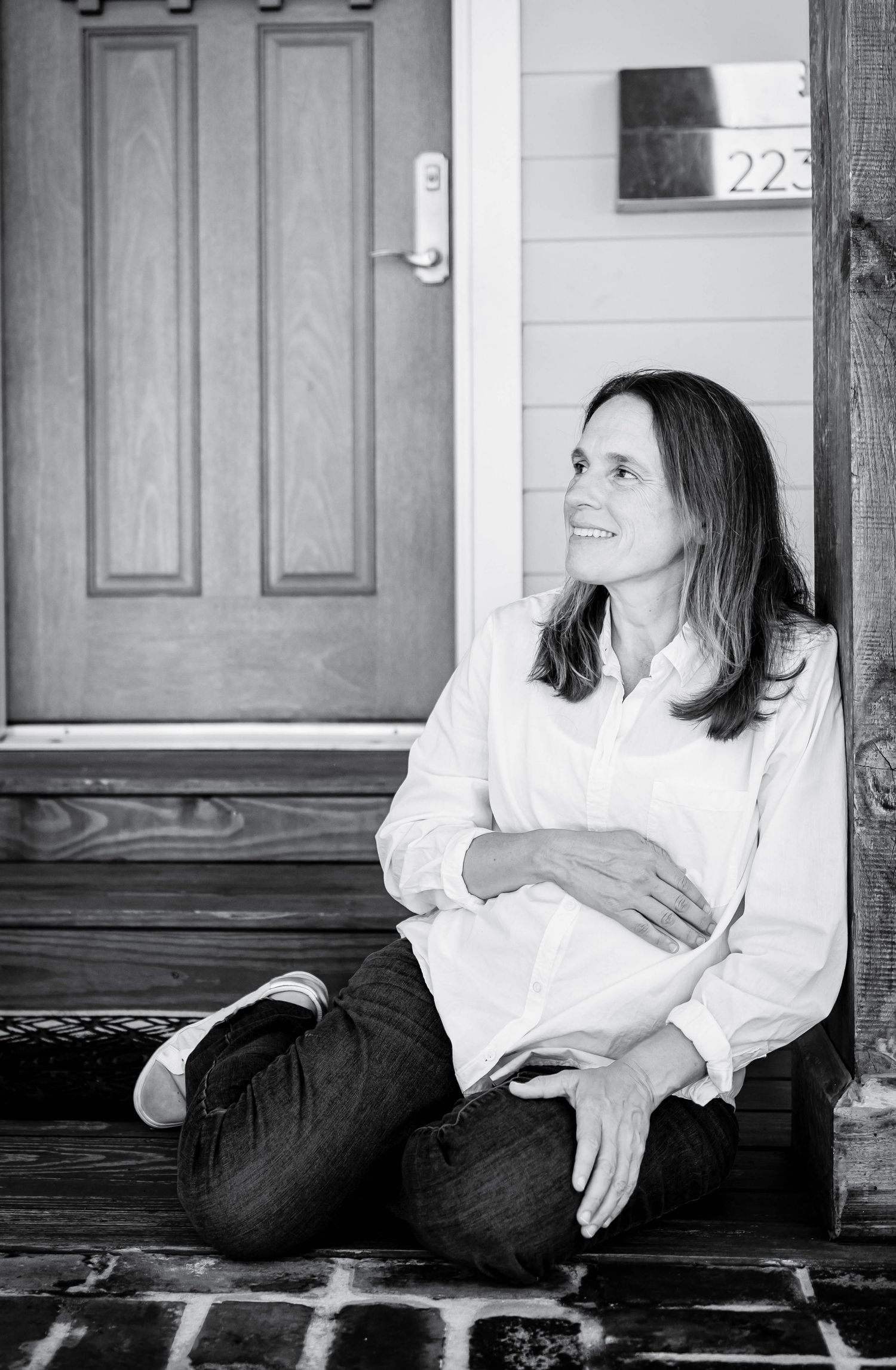Black and white photo of a smiling woman in white shirt and jeans sitting on a brick porch steps by a wooden door.
