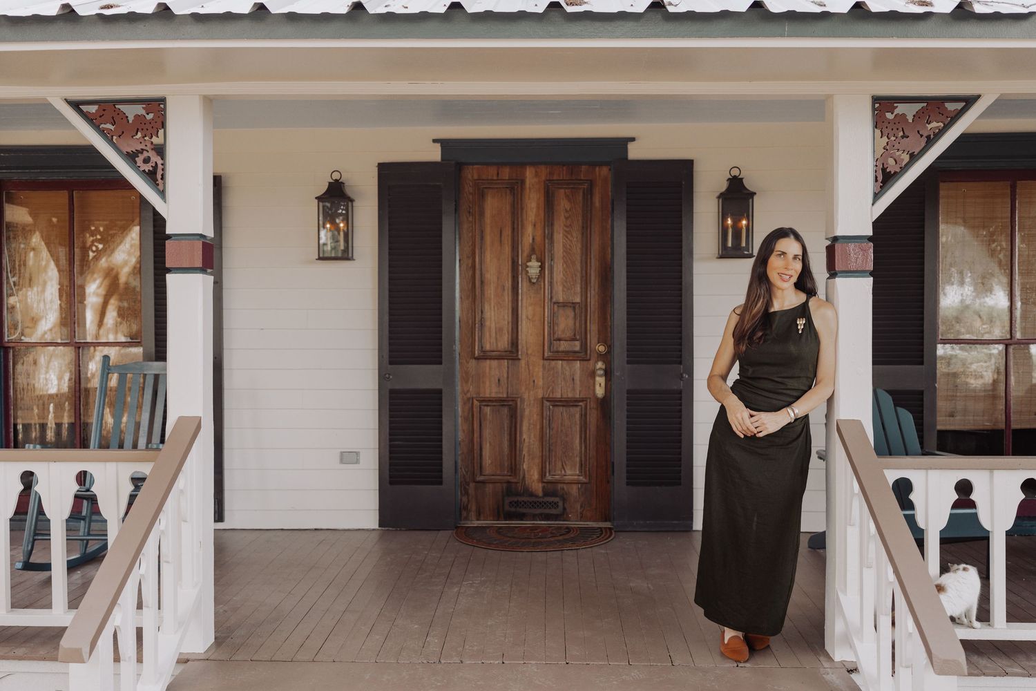 Woman in a long black dress stands on the front porch of a white farmhouse with a rustic wooden door and lanterns.
