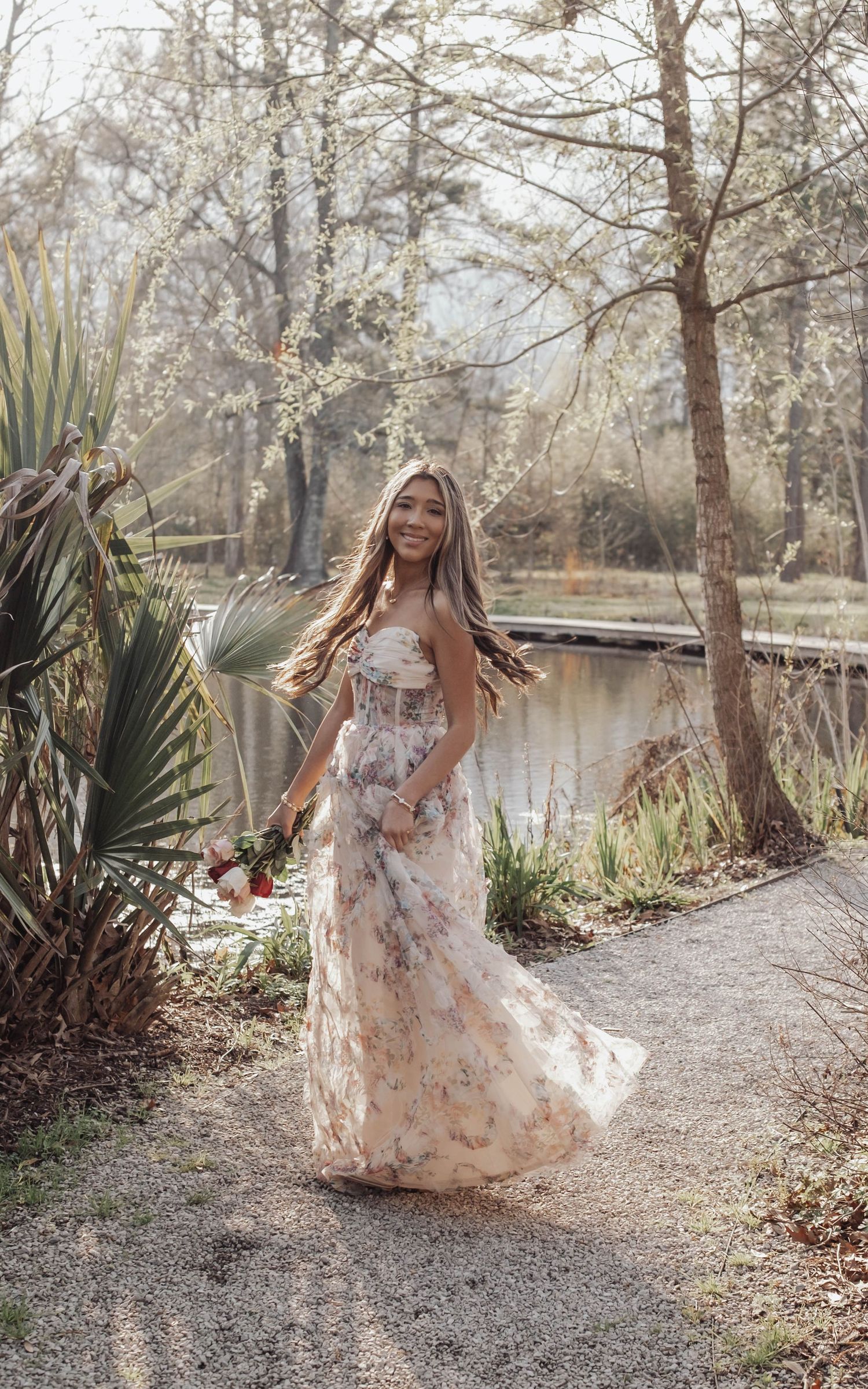 A floral maxi dress flows elegantly along a garden path with palm fronds and a serene pond in the background.