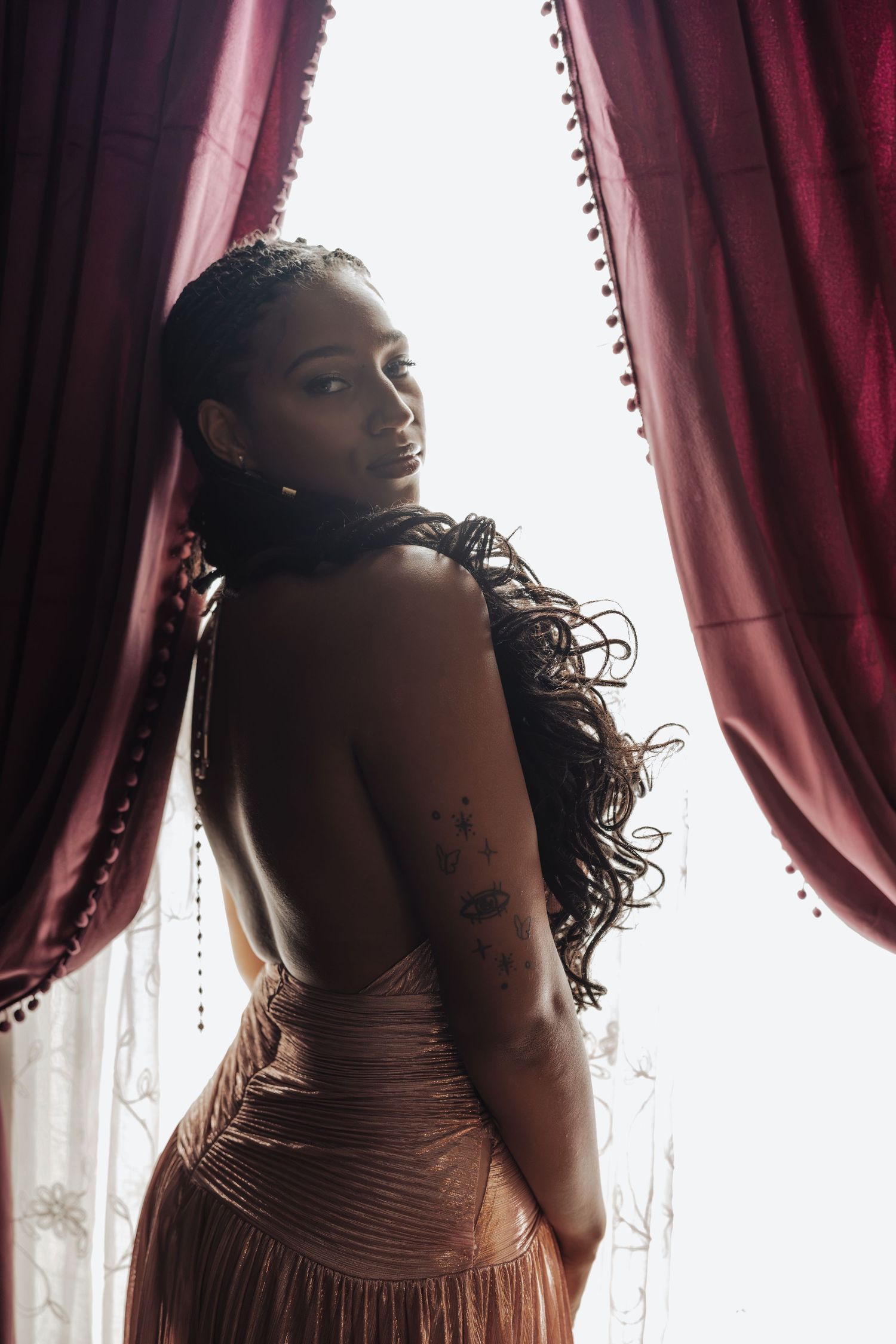 A woman in a backless bronze gown stands between red velvet curtains, glancing over her shoulder with long braided hair.