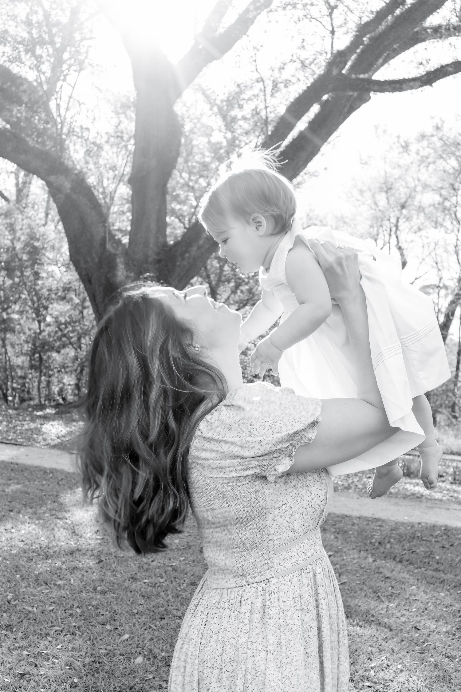 Black and white photo of a mother lifting her baby girl outdoors, sharing a joyful moment under sunlit trees.