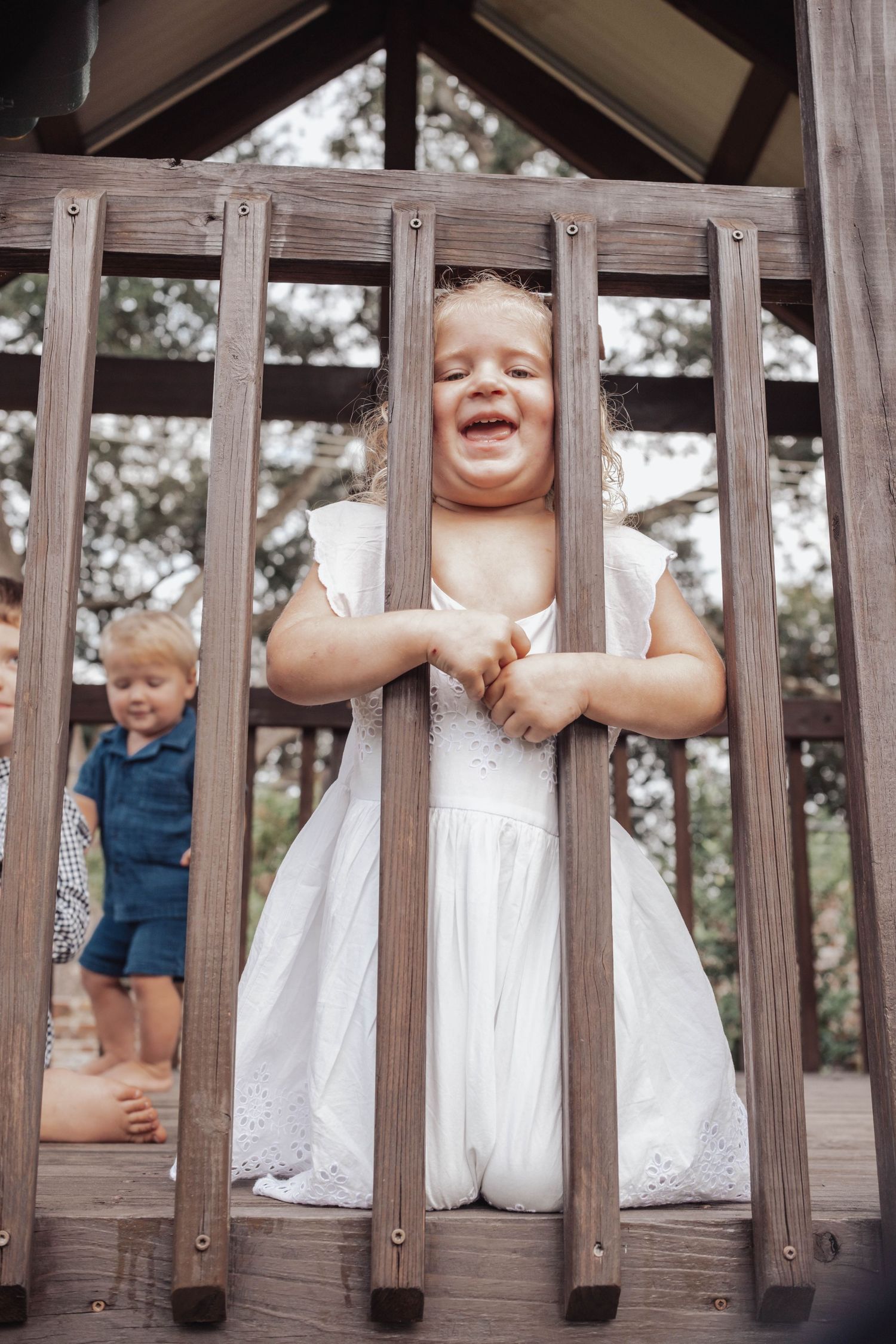 Three children in white dresses and blue overalls laugh joyfully while posing between wooden deck railings.