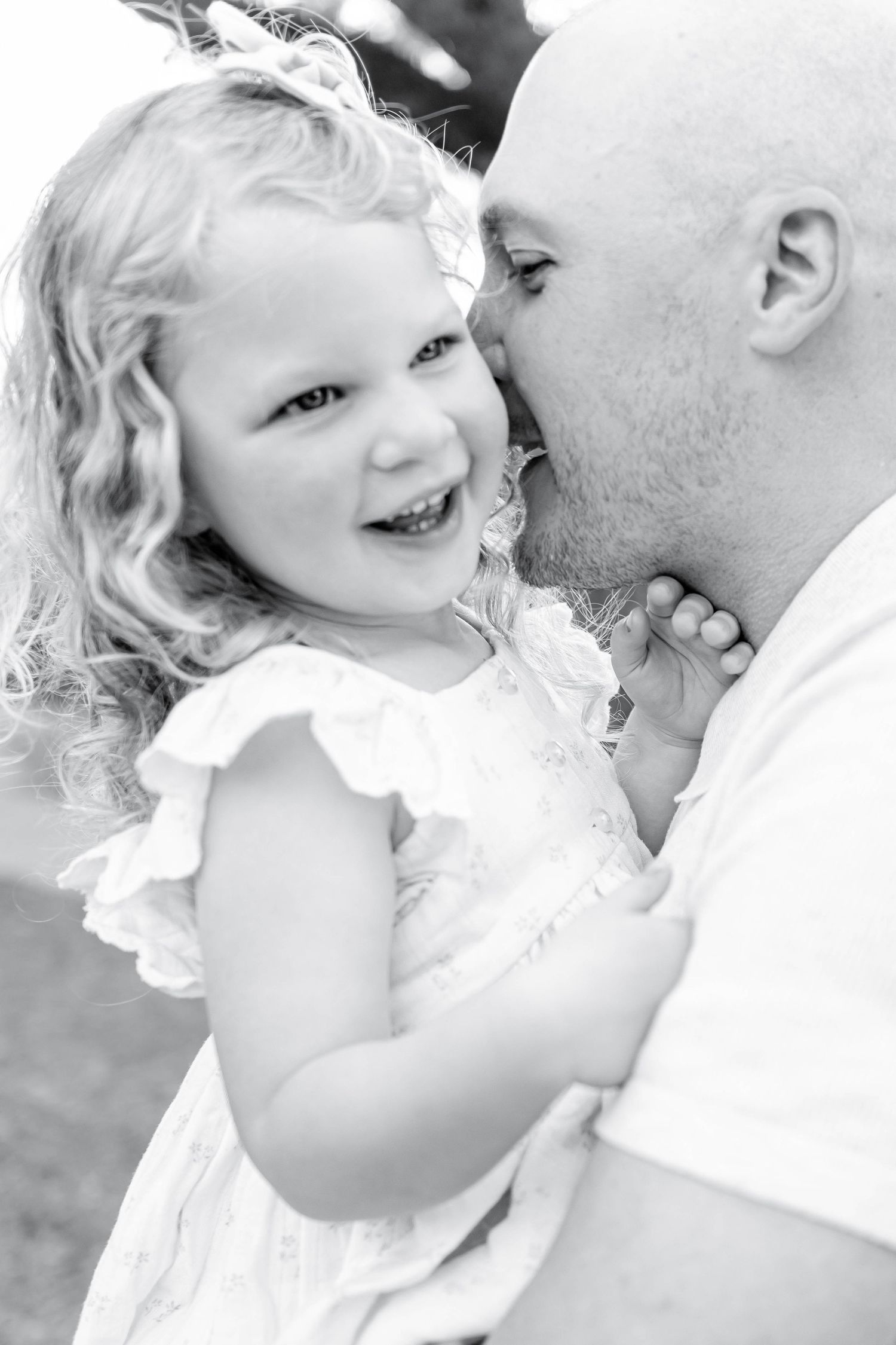 Black and white photo of a bald father kissing his laughing curly-haired little girl in a white dress.