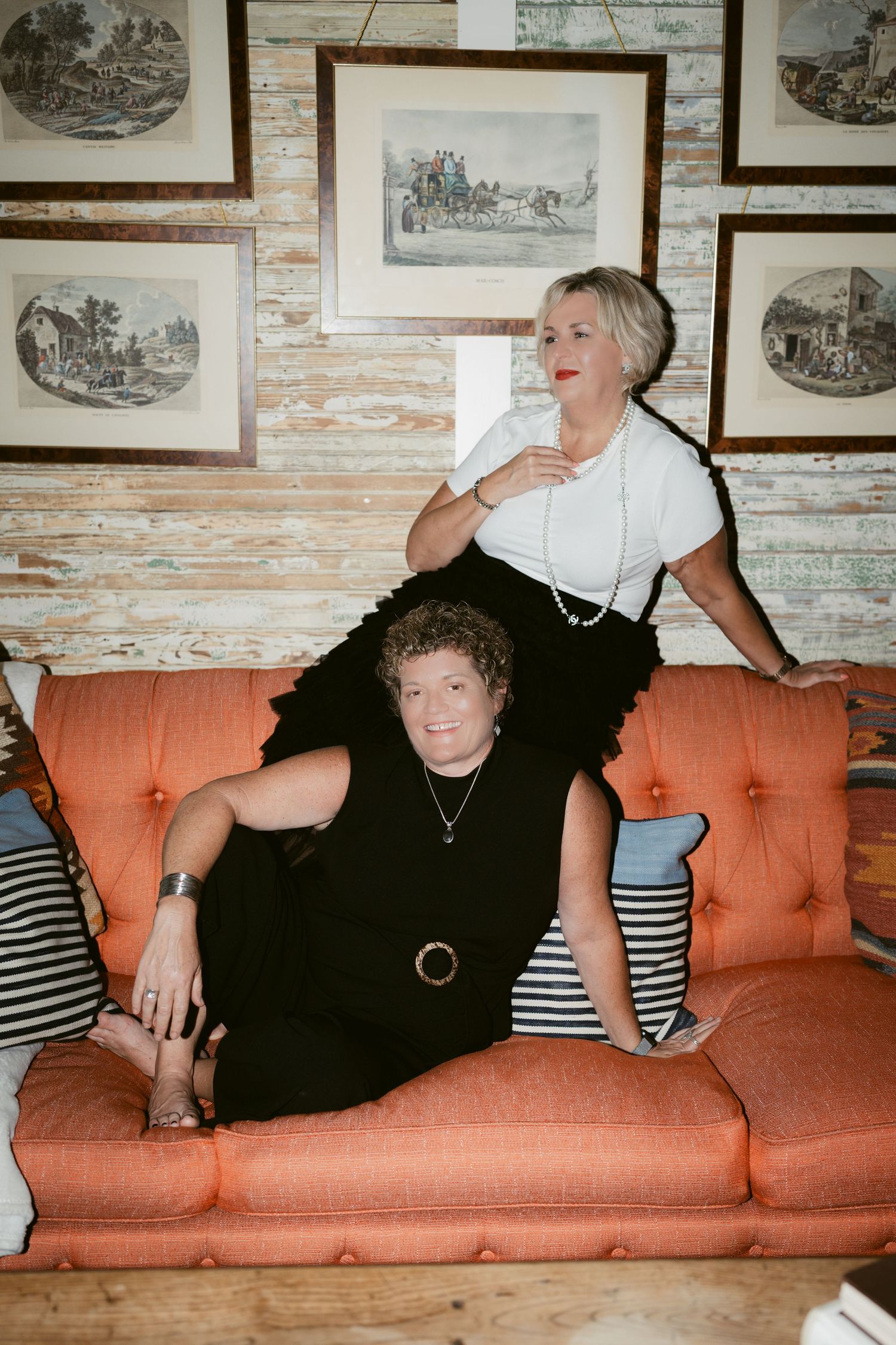 Two women in black outfits pose on an orange sofa, surrounded by framed artwork on a rustic white wall.