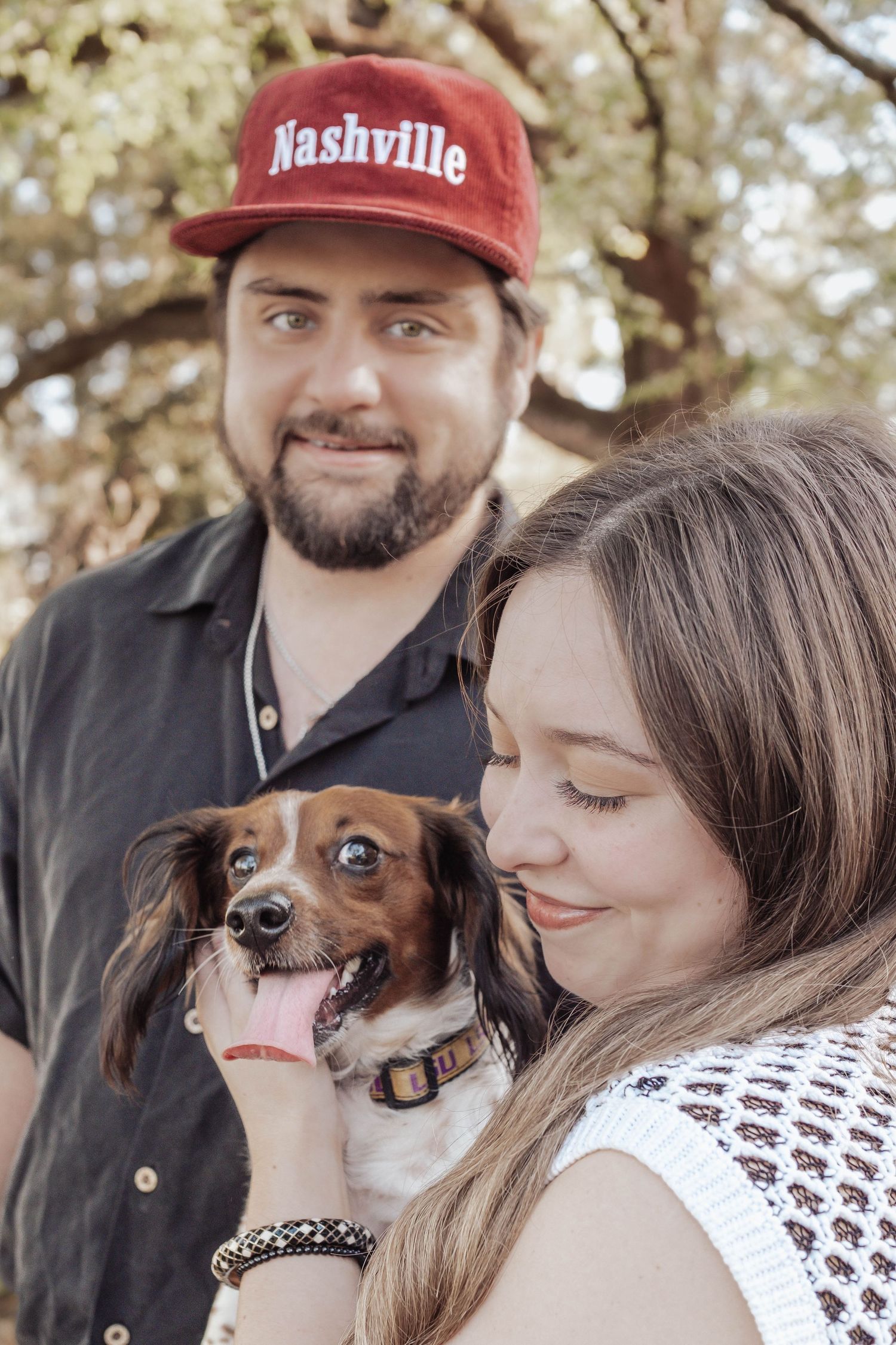 A couple with a small brown and white dachshund mix outdoors, man wearing a red Nashville cap.