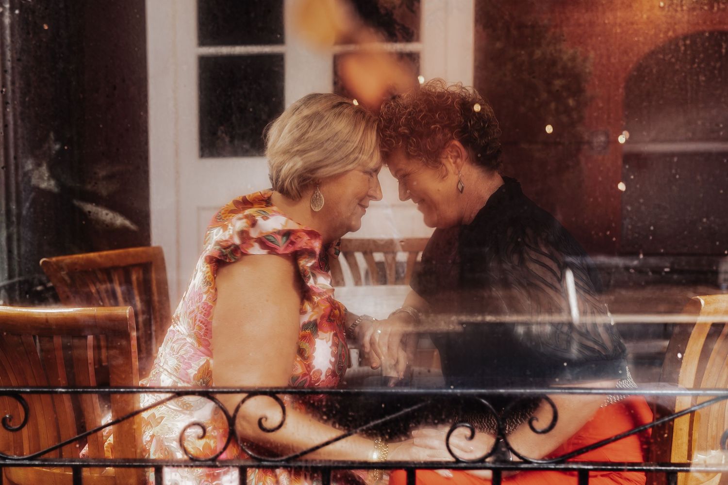 Two elderly women share an intimate moment together on a balcony, smiling and touching foreheads affectionately.