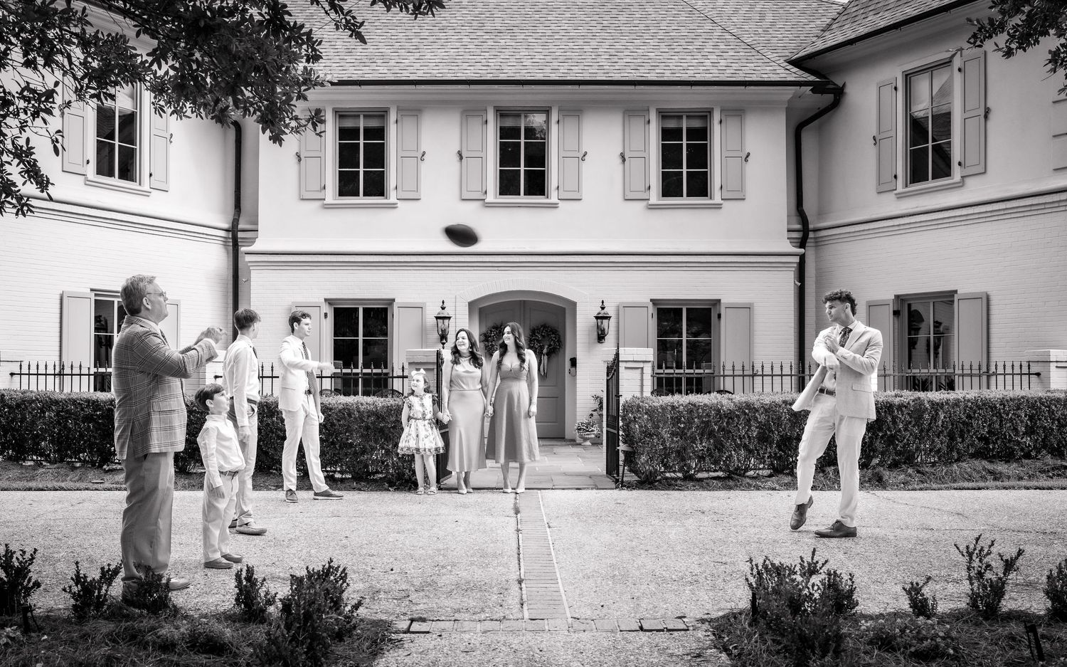 Black and white photo of elegantly dressed guests gathered outside a grand white estate home for an outdoor event.