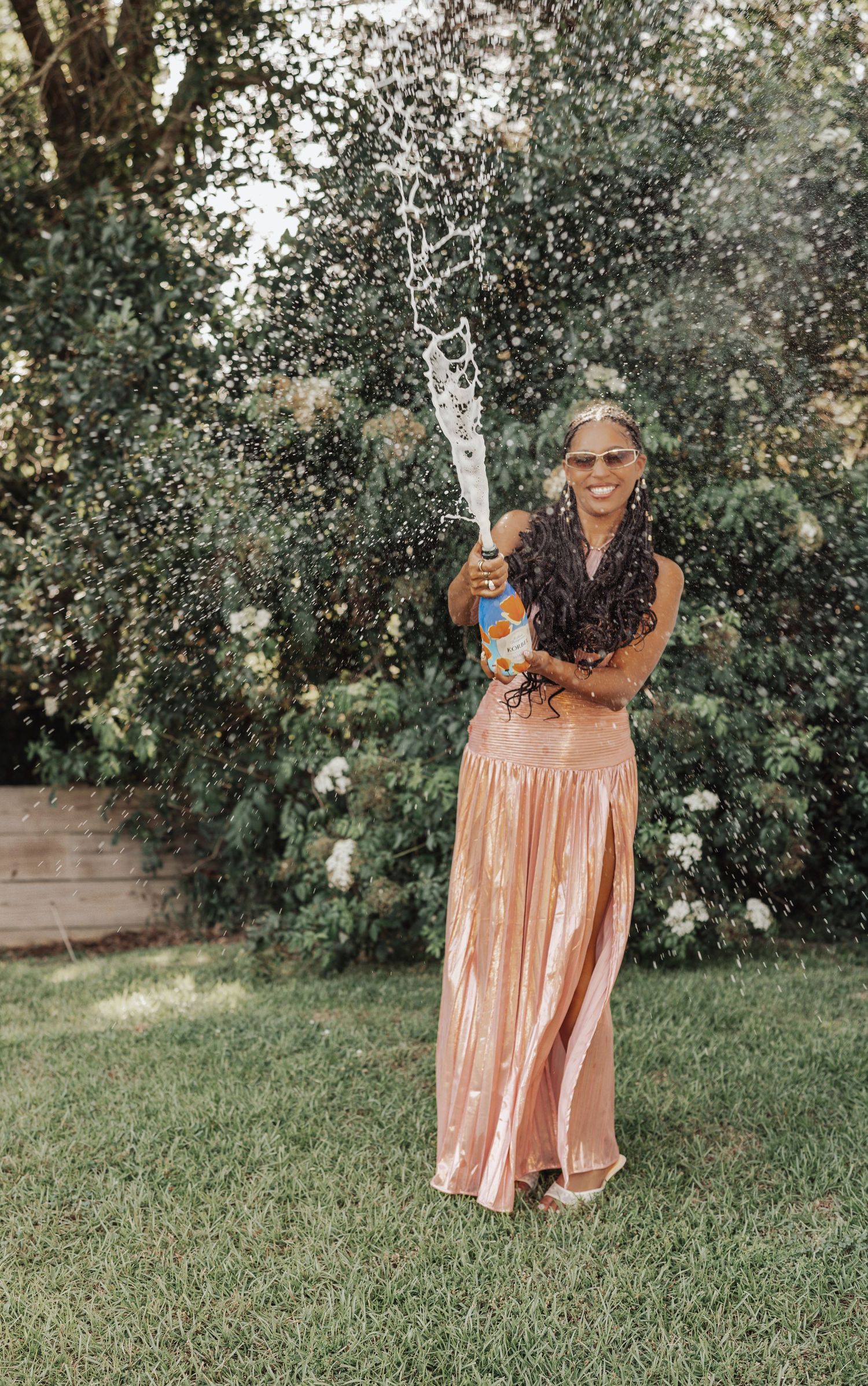 Woman in rose gold sequin skirt joyfully spraying champagne bottle in lush green backyard garden.